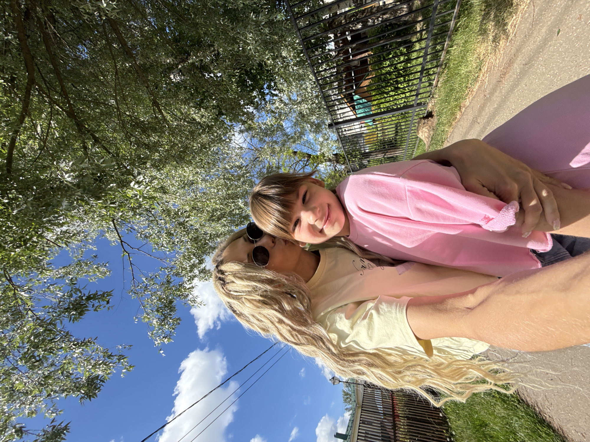 An outdoor selfie captures an affectionate moment between an adult woman and a young girl in Rusanovo, Russia, under a bright blue sky. The woman, with long, textured blonde hair and sunglasses perched on her head, wears a light yellow t-shirt and is embracing the girl. The girl, with light brown hair and bangs, smiles broadly at the camera while wearing a pink long-sleeved top.
The setting is a sunny day, with lush green trees and foliage providing a natural canopy overhead, casting dappled light. White, fluffy clouds are scattered across the clear blue sky. In the background, a black metal fence runs horizontally, separating a grassy area from a paved path or road. Hints of distant structures are visible beyond the fence, and a utility wire crosses the sky. The overall mood is joyful and warm, emphasizing the close bond between the two individuals. No visible text is present in the image.
