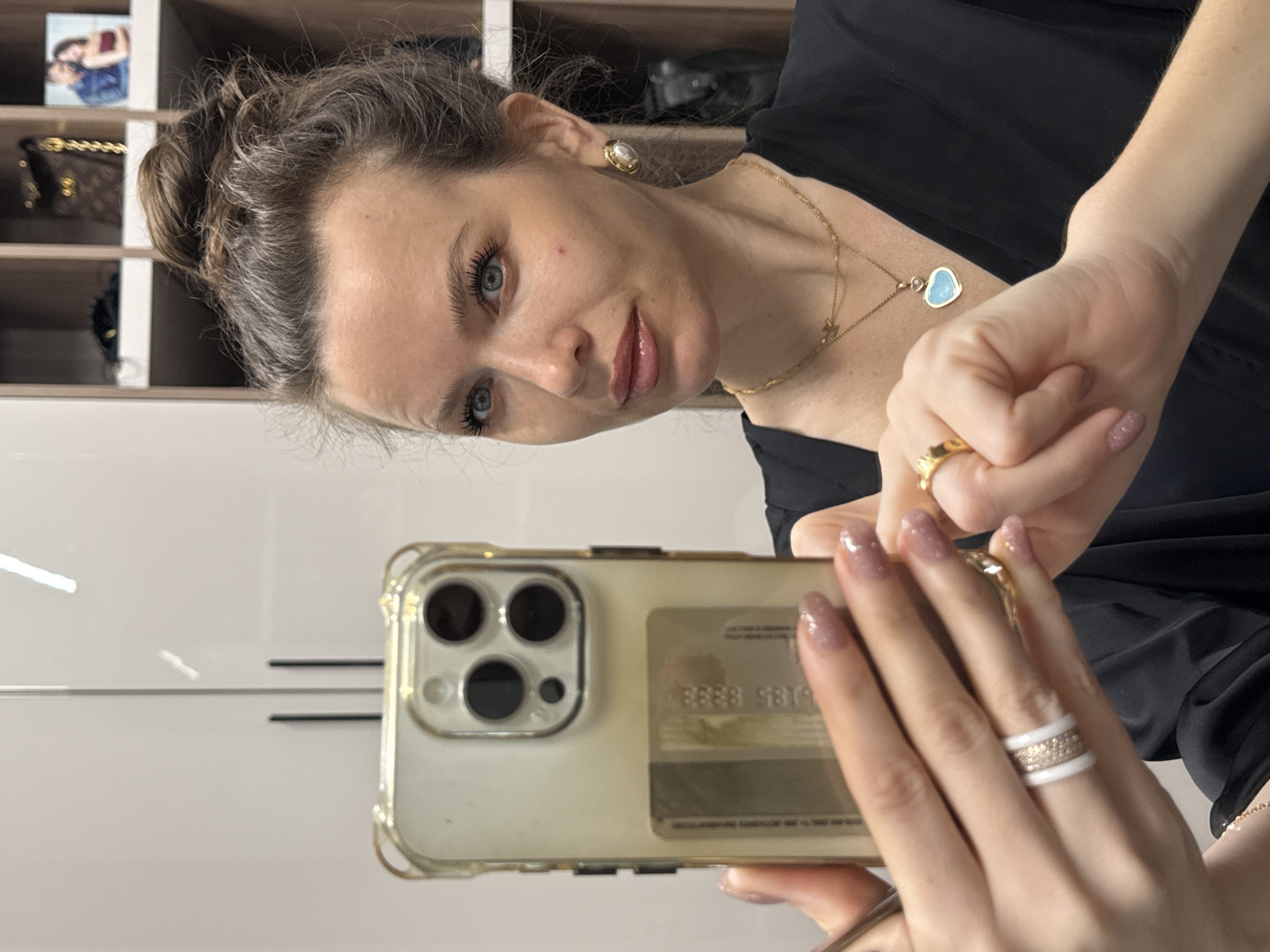A woman is taking a selfie with her smartphone. She has light skin, blue eyes, and her hair is pulled back. She is wearing gold jewelry: earrings, a necklace with a blue heart pendant, and rings on both hands. The phone has a clear case and a credit card attached to the back. The background includes a shelf with various items and a glossy white wall. The location is in Khimki, Russia.