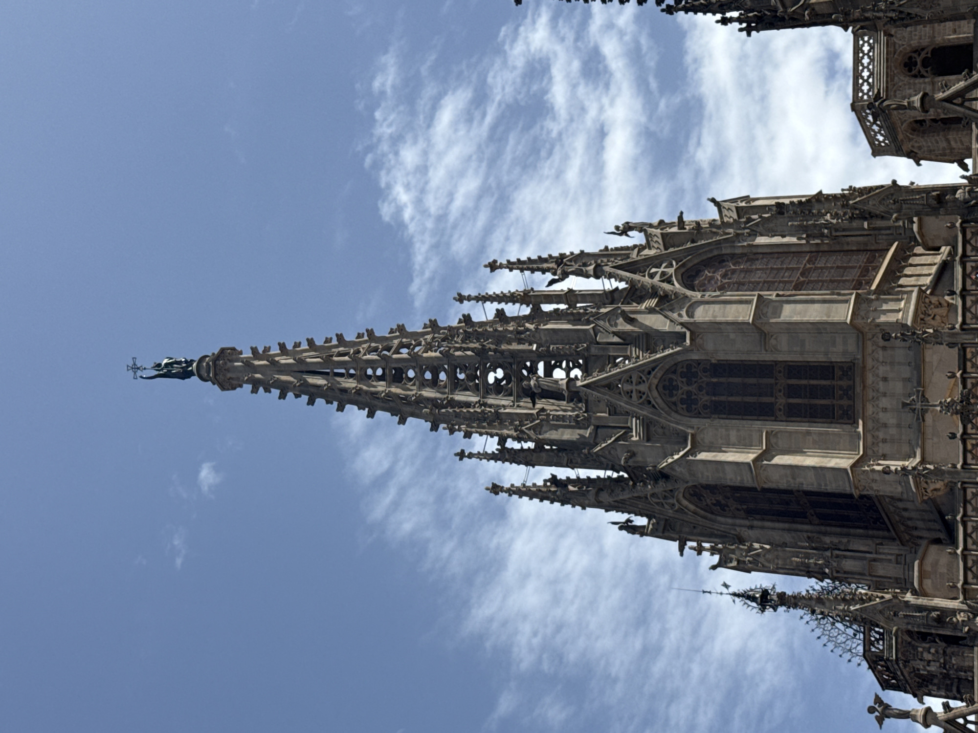 The image shows a low-angle, close-up view of the spires and upper facade of the Barcelona Cathedral, also known as the Cathedral of the Holy Cross and Saint Eulalia, located in the Gothic Quarter of Barcelona, Spain. The architecture is characterized by Gothic Revival style, with intricate stone carvings, pointed arches, and ornate spires reaching towards a bright blue sky dotted with wispy white clouds. A statue of a figure holding a cross stands atop the tallest central spire. Gargoyles are visible adorning the building's ledges. The stone appears weathered, suggesting the age of the structure. No people are visible in the frame.