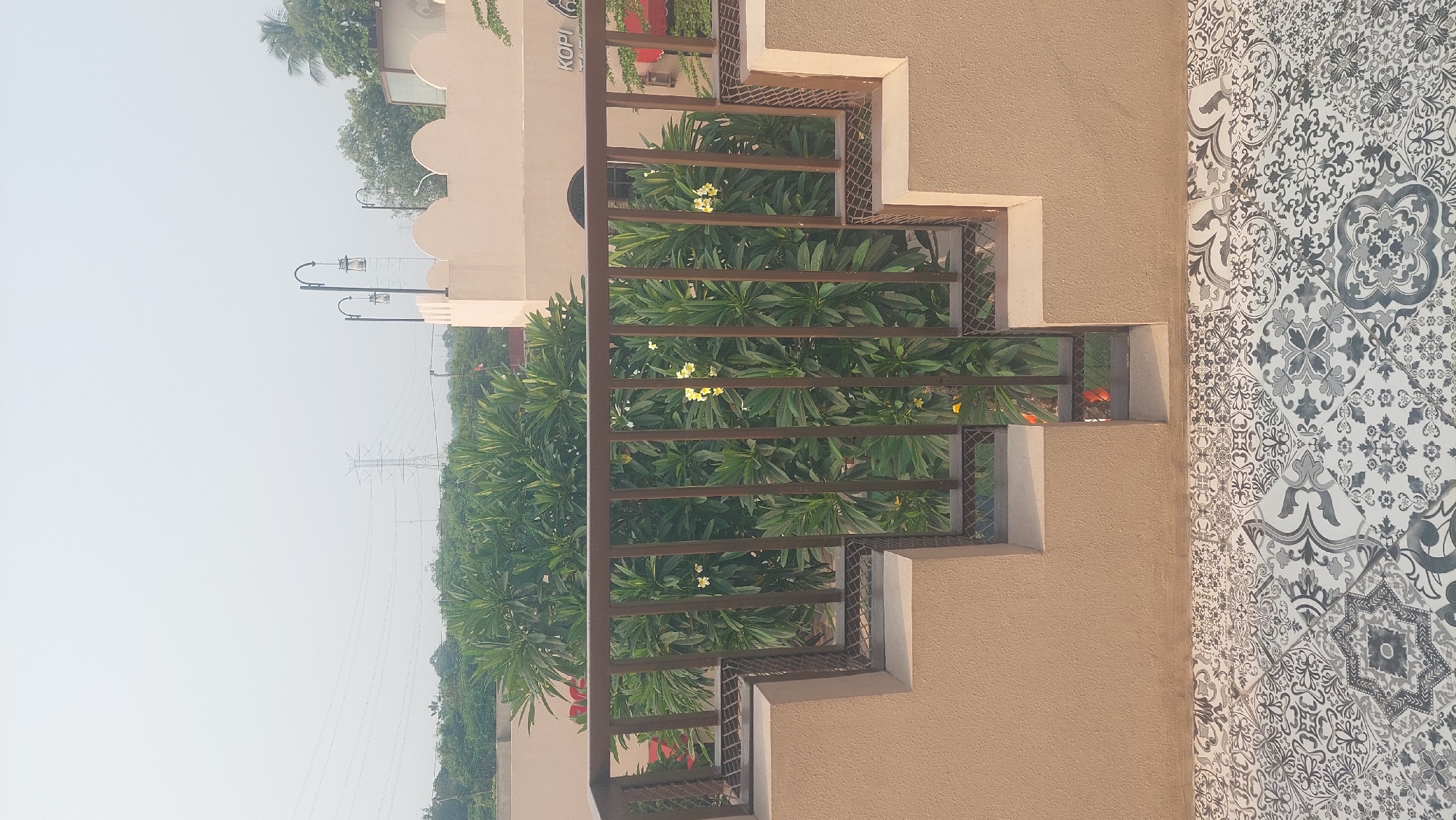 In Cileungsi, Indonesia, the image captures a serene outdoor scene, possibly a cafe or resort. In the foreground, a patterned tiled floor leads to a beige, stepped wall and a brown metal railing. Behind the railing, lush green trees with small yellow flowers are visible. In the background, a beige building with decorative cutouts and arched windows features the text "KOPI" on its facade. The sky is overcast, and power lines and a transmission tower are visible in the distance, suggesting an urban or semi-urban environment. The scene appears peaceful and well-maintained.