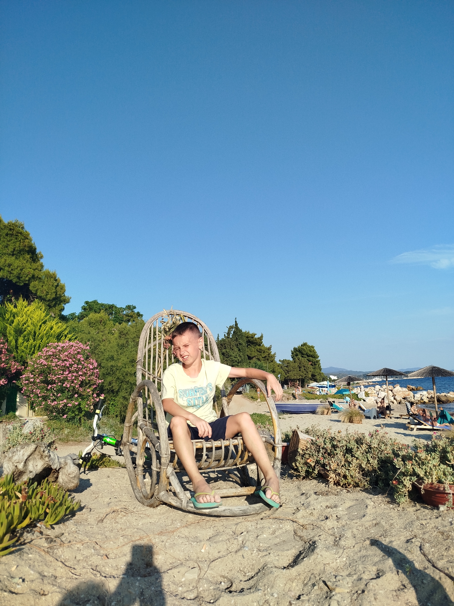 A young boy with light brown hair and fair skin is sitting in a round, wicker chair on a sandy beach. He is wearing a light yellow t-shirt with blue lettering that reads "SURFER'S STYLE" and dark blue shorts. He has on green flip-flops. His eyes are squinting and his mouth is open in a smile. The chair is made of woven wood and has large, round sides that resemble wheels. Behind the boy, there are trees and bushes, with pink flowers on a bush to the left. In the background, there is a beach with people relaxing under umbrellas, lounge chairs, and the sea. The sky is clear and blue. The overall scene suggests a sunny day at a beach resort in Sithonia, Greece.