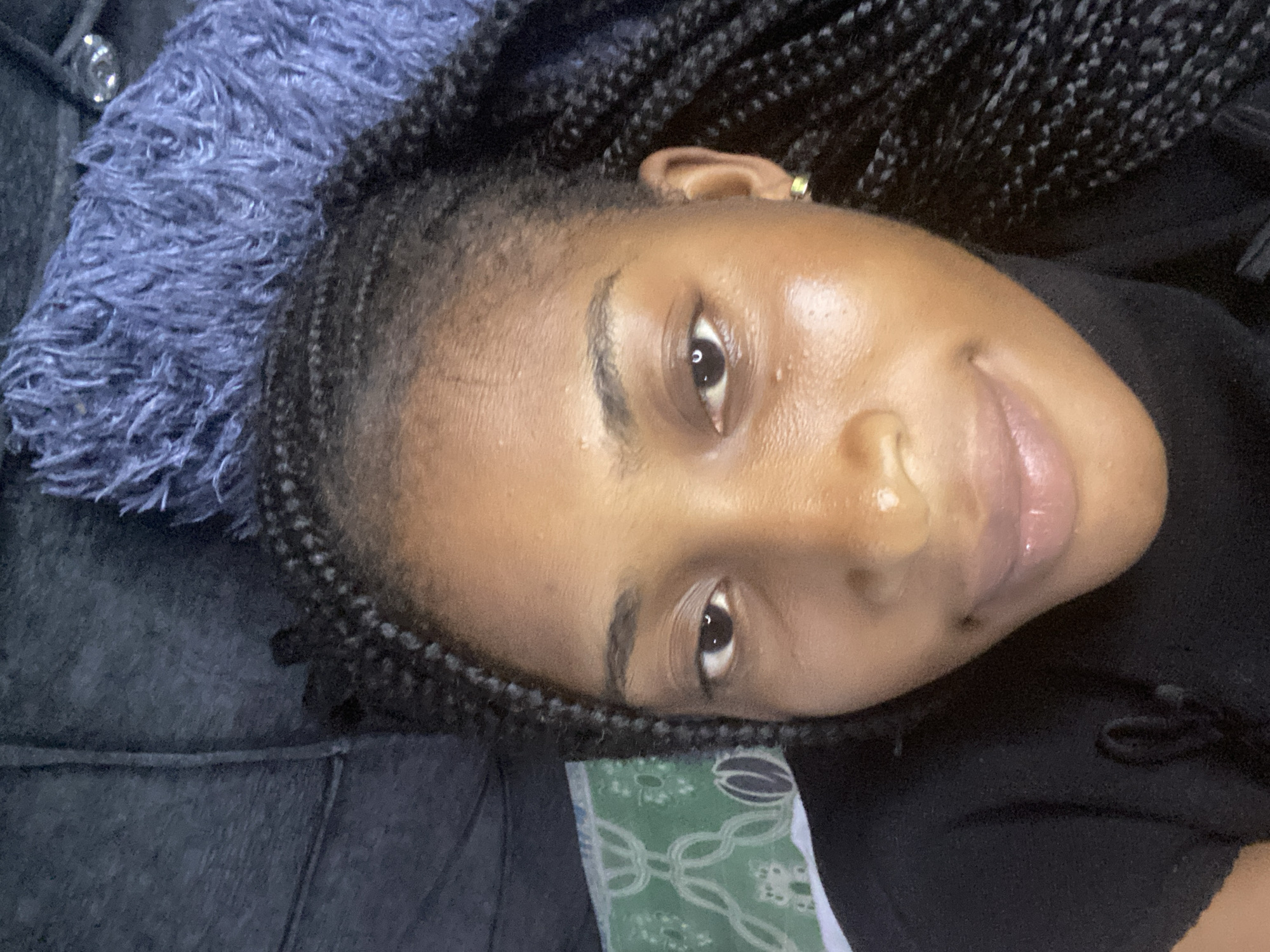 A close-up, overhead, selfie-style shot shows a young Black woman with braided hair lying down, looking towards the camera. She is wearing a black turtleneck. A textured blue throw is visible behind her head, and a dark gray sofa is to her left. A patterned green fabric is under her left side. She appears to be in an indoor setting, possibly a living room, in Kubwa, Nigeria.