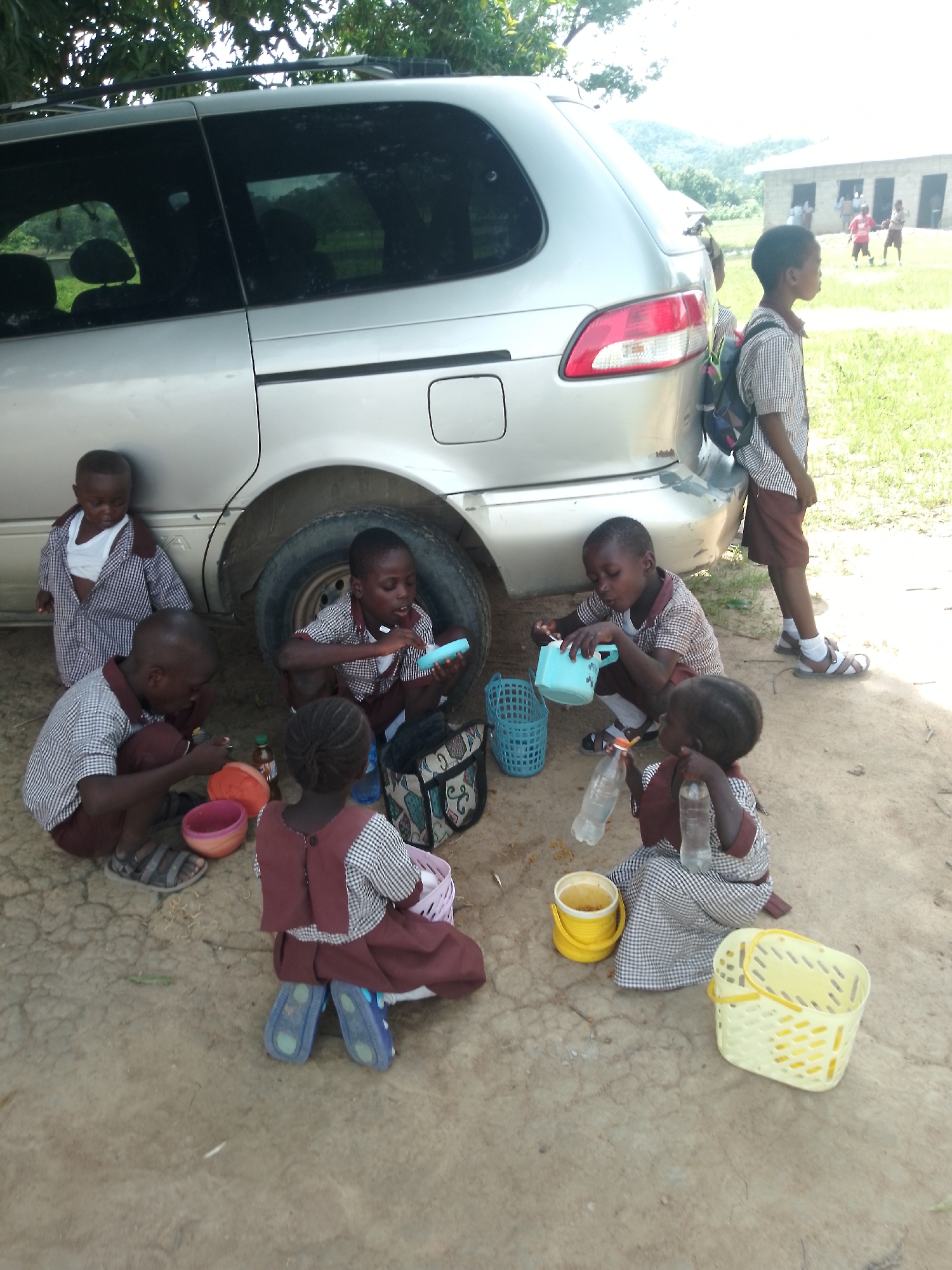 In Serti, Nigeria, a silver minivan is parked on a dirt road under the shade of trees. A group of five young children, dressed in school uniforms consisting of plaid shirts and maroon shorts or skirts, are gathered around the vehicle. They appear to be having a break or meal. Two of the children are drinking from plastic bottles. One child holds a light blue plastic cup, and another is holding a similar object, possibly a toy. There are various items scattered on the ground including a red bowl, a yellow bucket, and a yellow plastic basket. In the background, another child in a school uniform stands by the minivan, and further in the distance, a school building is visible with other children playing outside. The ground is dry and cracked, indicating a rural or semi-rural environment.