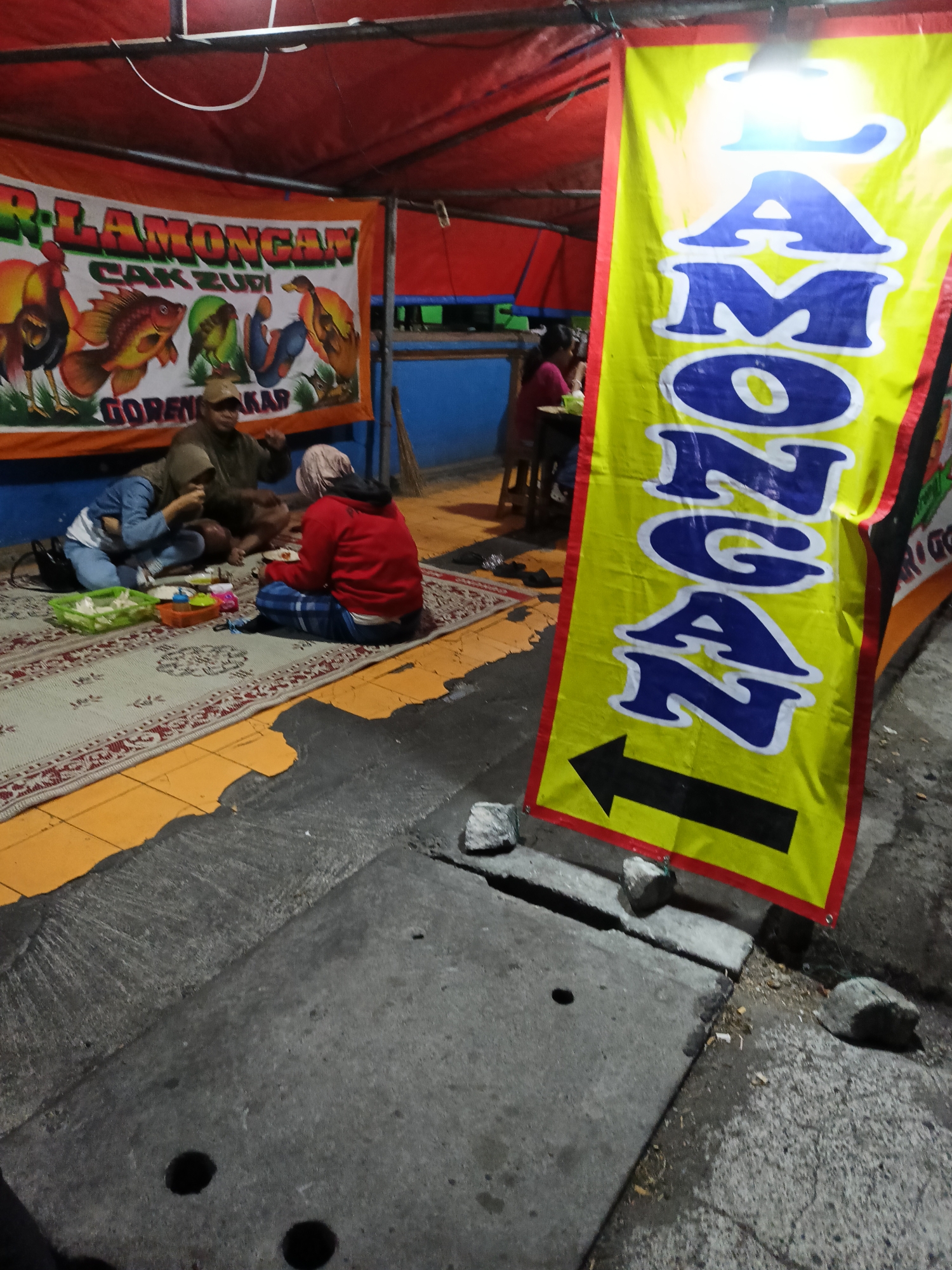 The image depicts an outdoor scene at night, likely a food stall in Kebondalem Kidul, Indonesia. A red tent provides overhead cover. To the left, a banner displays a rooster, a goldfish, and ducks with the text "R-LAMONGAN CAK ZUDI GORENG AKAR." In the foreground, three individuals are seated on a patterned rug, engaged in eating. One person, wearing a red jacket and blue pants, is facing away from the camera, while another in a blue denim jacket and jeans is interacting with someone who is partially obscured. A man in a brown jacket and cap is also present. To the right, a large yellow banner with red trim features the word "AMONGAN" in large blue letters, followed by a black arrow pointing to the left. The ground consists of concrete and asphalt, with some stones scattered around. The overall impression is of a casual, open-air dining setting.