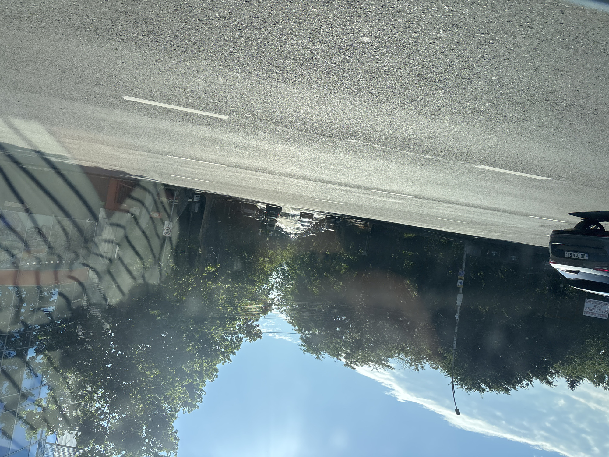 The image depicts a street scene in Tbilisi, Georgia, on a bright, sunny day. The perspective is from inside a vehicle, looking out at the road ahead. The road is asphalt with white lane markings. On the left side of the road, a dark-colored SUV with the license plate "JG-014-GJ" is visible, along with trees and a lamppost. Further down the road, a line of cars is seen moving forward. On the right side, a modern building with reflective glass windows and trees in front of it is partially visible. The sky is blue with scattered white clouds, and the sun is shining brightly, creating lens flare. The overall impression is of a typical urban environment with traffic and vegetation under clear skies.