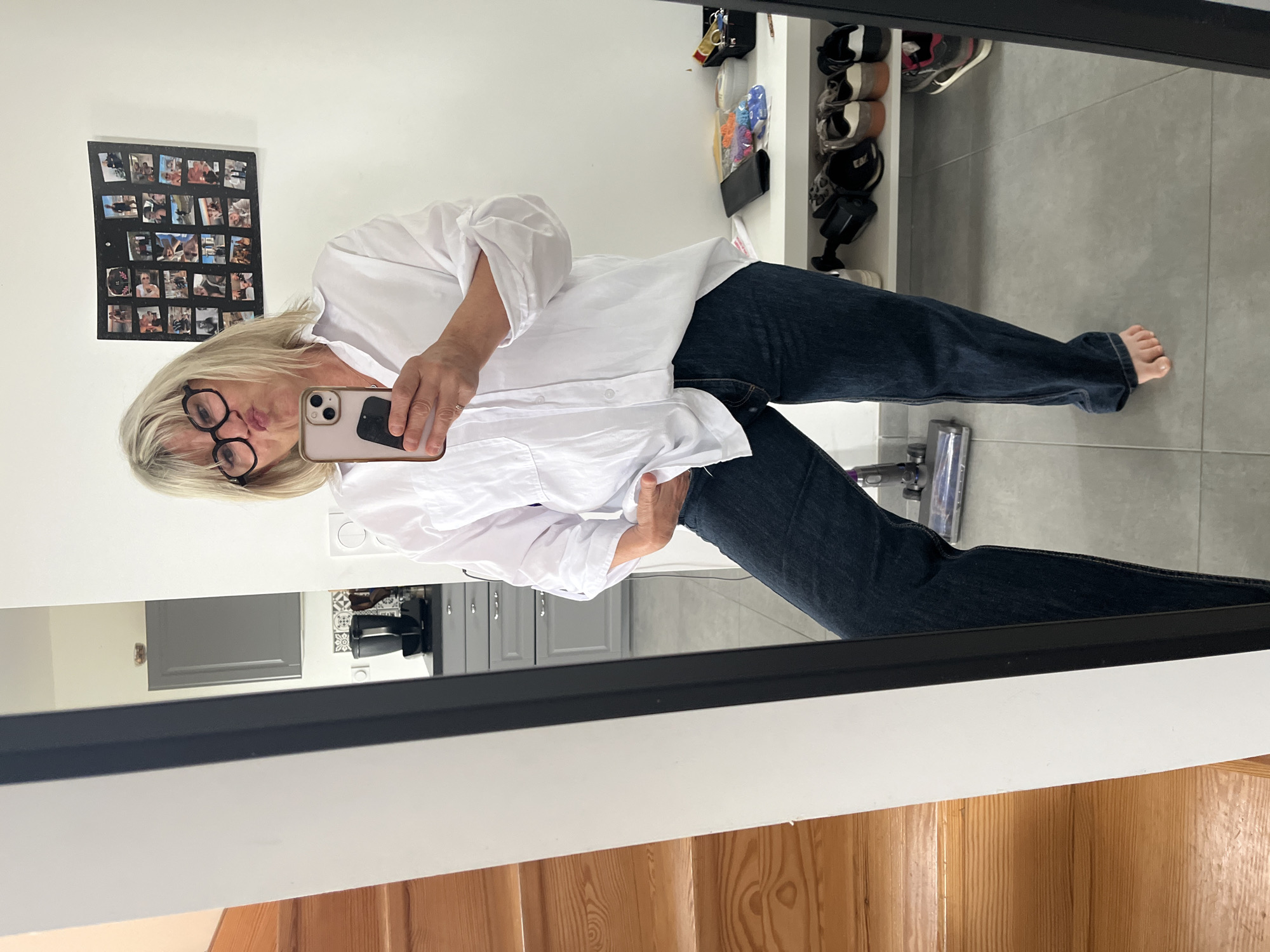A woman is taking a selfie in a mirror. She is wearing a white blouse, dark jeans, and black framed glasses. She has short blonde hair. The background includes a tiled floor, a shoe rack, a black framed photo board on the wall, kitchen cabinets, stairs, and a vacuum cleaner. The location is Chambœuf, France.
