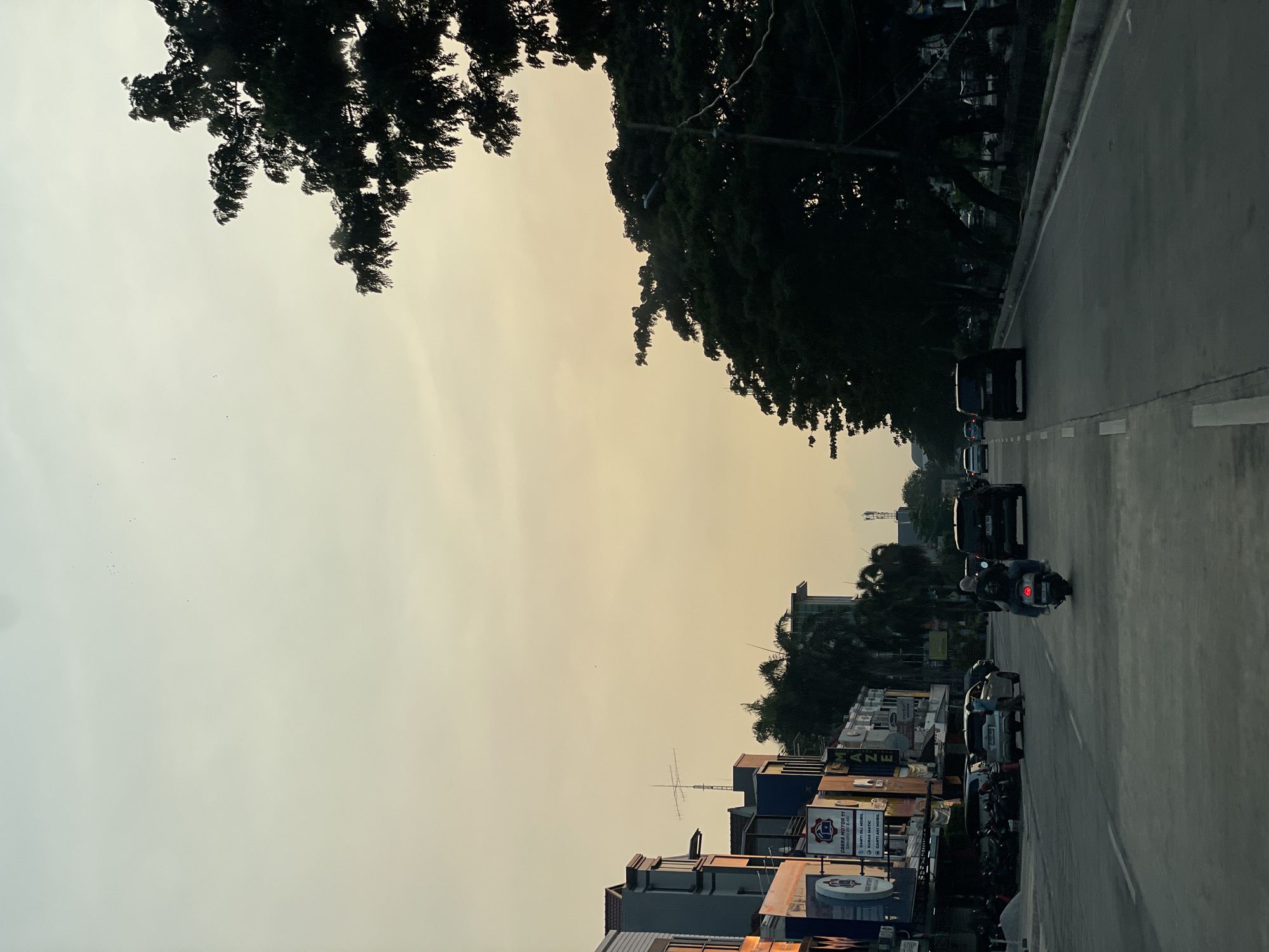 The image shows a street scene in North Jakarta, Indonesia. The photo is taken at ground level, depicting a multi-lane asphalt road with vehicles, including cars and a motorcycle, moving along it.
The sky is overcast, with a yellowish-grey hue suggesting possible haze or dusk. Trees are visible lining both sides of the street, with dense foliage. Buildings and commercial establishments are present on one side of the road, identifiable by various signage.
No people can be clearly identified other than the motorcyclist on the road. The scene suggests an urban environment with typical transportation and infrastructure.
