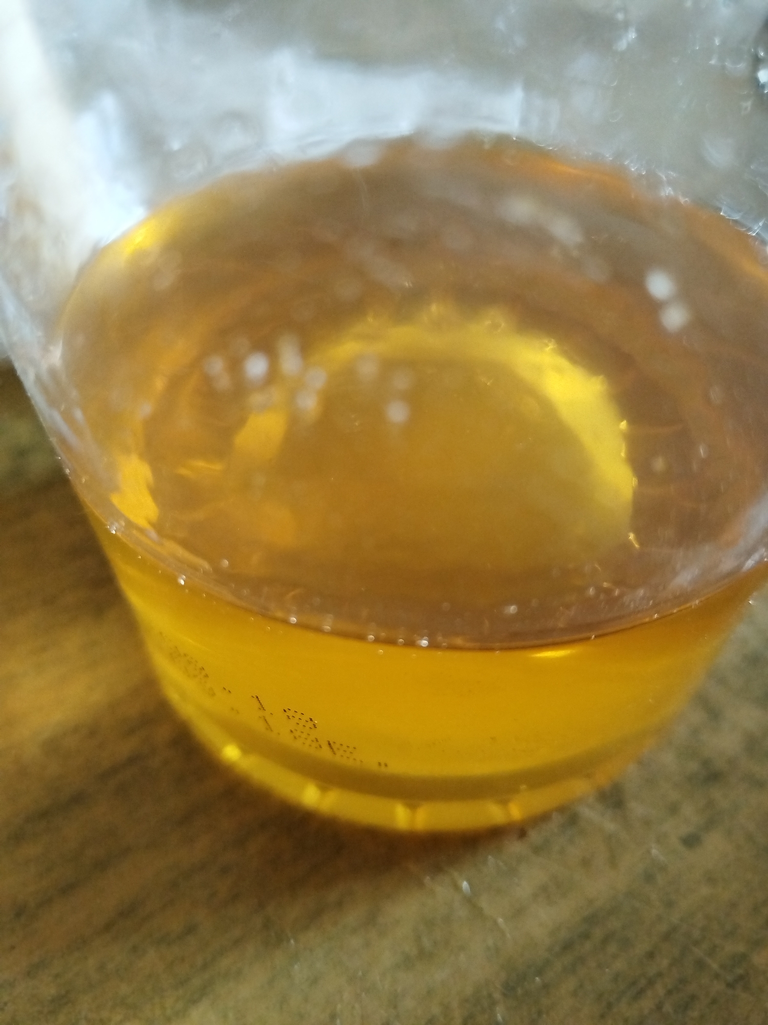 A close-up view shows a clear glass containing a golden-colored liquid, likely a beverage such as beer or juice, resting on a wooden surface. The liquid shows some small bubbles clinging to the inside of the glass. In the lower portion, small embossed numbers and letters on the glass are partially visible. The environment is out of focus. There is no information to determine that the photo was taken in Chaikovsky, Russia.