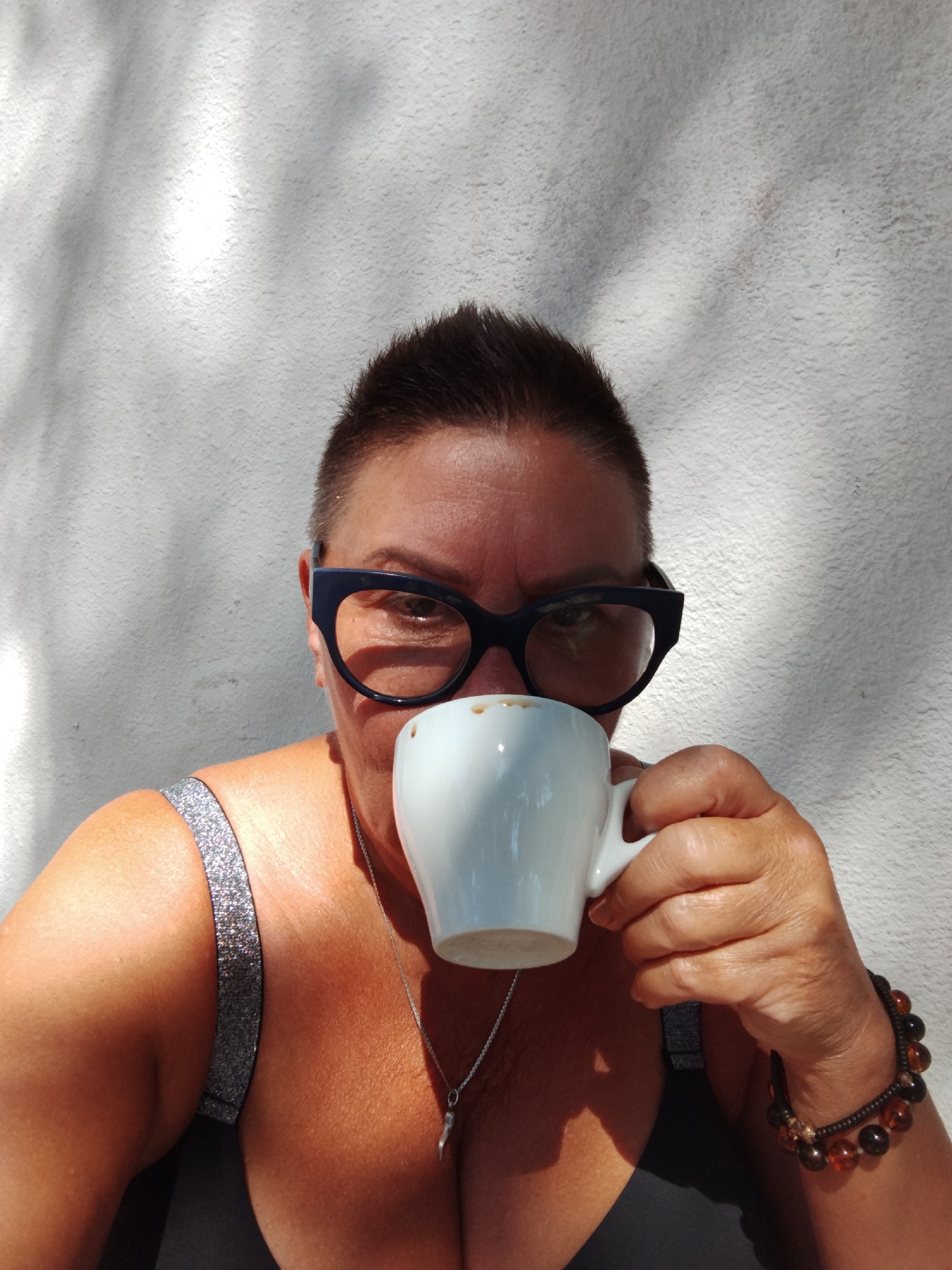 A medium shot captures a woman with a short, dark buzz cut and dark-rimmed glasses looking directly at the camera while holding a white espresso cup to her lips. She is wearing a dark top with glittery straps. Her right hand, adorned with two beaded bracelets, is holding the cup. The background is a textured white wall, partially obscured by dappled light and shadows, suggestive of an outdoor setting. The image appears to have been taken on a sunny day, possibly in Peschici, Italy, given the typical Mediterranean ambiance suggested by the lighting and the wall texture. The woman's gaze is direct, and her expression is neutral. There are no other individuals visible in the frame. The overall scene is simple and focused on the woman and her interaction with the coffee cup.