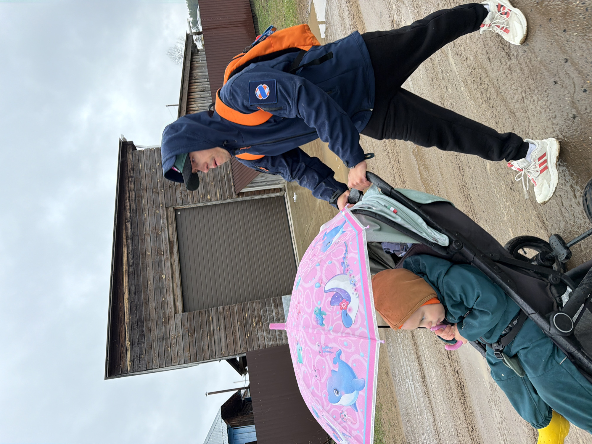 An adult is pushing a child in a stroller along a wet, unpaved road in Chaikovsky, Russia. The sky is overcast.
The adult, appearing male, is on the right side of the image, facing partially left, and is dressed in a navy blue hooded jacket with an orange backpack. The jacket has a circular logo on the sleeve, featuring concentric rings in orange, blue, and white. He wears black pants and white sneakers with red Adidas stripes. A black baseball cap is visible under his hood. He is holding the handle of a black stroller.
Seated in the stroller on the left is a young child, wearing a dark green jumpsuit or overalls, a brown or orange beanie, and bright yellow boots. The child is holding a pink umbrella adorned with marine animals like dolphins, jellyfish, starfish, and a turtle. The child's face is mostly obscured but appears calm, looking downwards.
The setting features several simple wooden buildings in the background, suggesting a rural or semi-rural environment. One building has vertical wooden planks, while another has horizontal corrugated metal siding. The ground is wet and muddy, indicating recent rain or damp conditions. The overall atmosphere is outdoors during an overcast daytime. No distinct text other than the Adidas stripes is clearly legible.