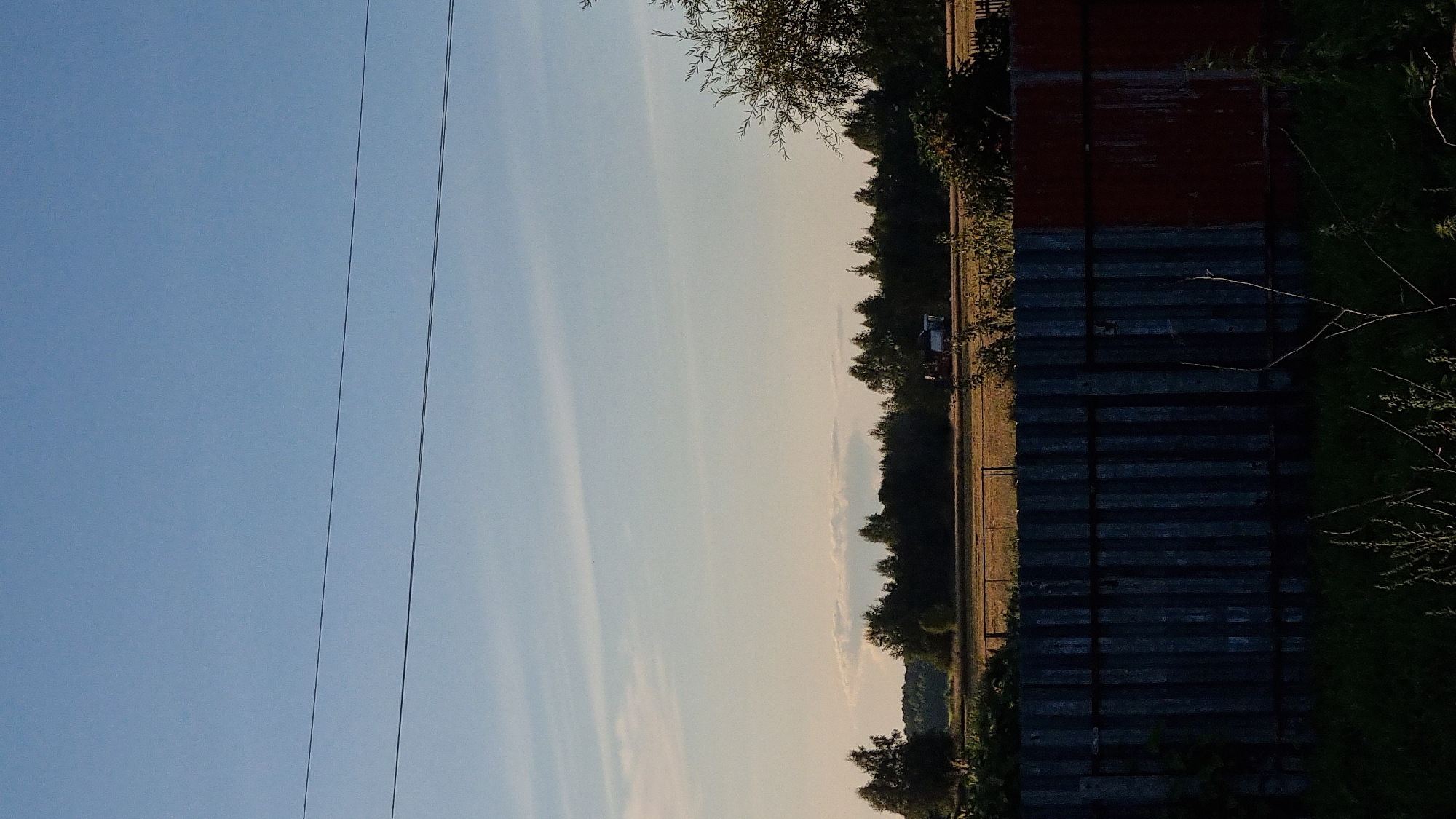 The image depicts a rural landscape in Vysokoe, Russia, during the late afternoon or early evening. In the foreground, a dark, corrugated metal fence runs horizontally across the frame. The fence is partially covered in vegetation. Beyond the fence, a field of harvested crops, likely grain, is visible. A line of trees forms a dense border at the far edge of the field, with a forest extending behind them. A red and white combine harvester is operating in the field. The sky is a clear, pale blue with thin, wispy clouds, suggesting a clear day. Two power lines are stretched across the upper portion of the image. The overall impression is one of a working agricultural landscape under a tranquil sky.