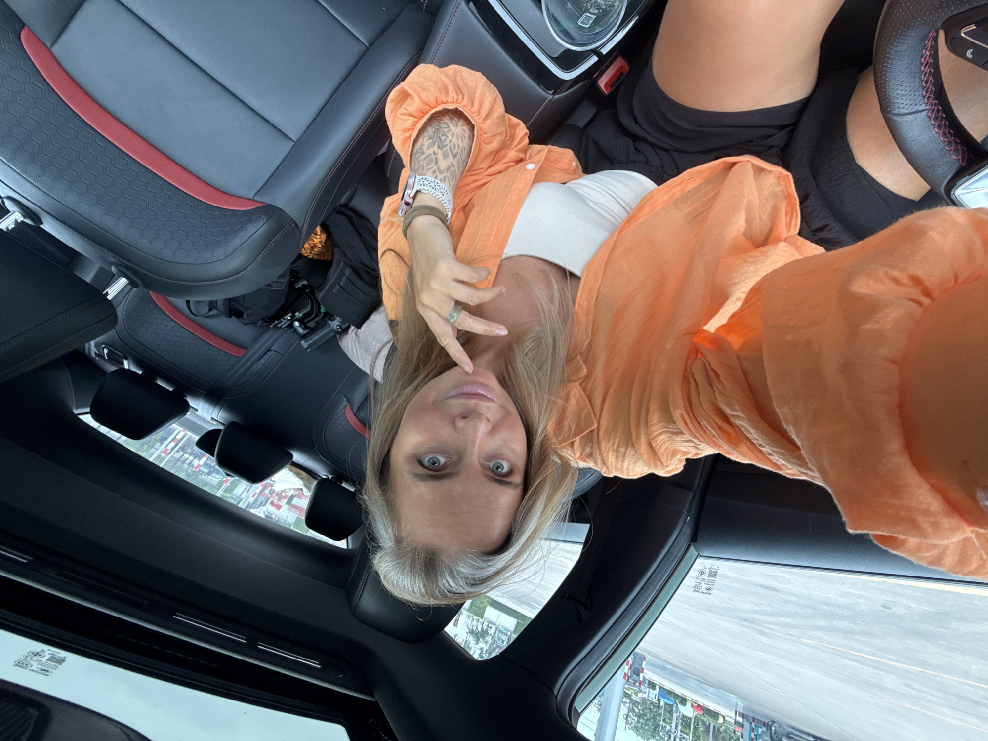 A woman with blonde hair and blue eyes is taking a selfie in the driver's seat of a car. She is wearing an orange shirt, a white tank top, and black shorts. The car appears to be parked outside in what looks like a busy area with buildings and vehicles visible in the background. The interior of the car is visible, showing black leather seats with red accents and a sunroof. The woman is touching her chin with her right hand.