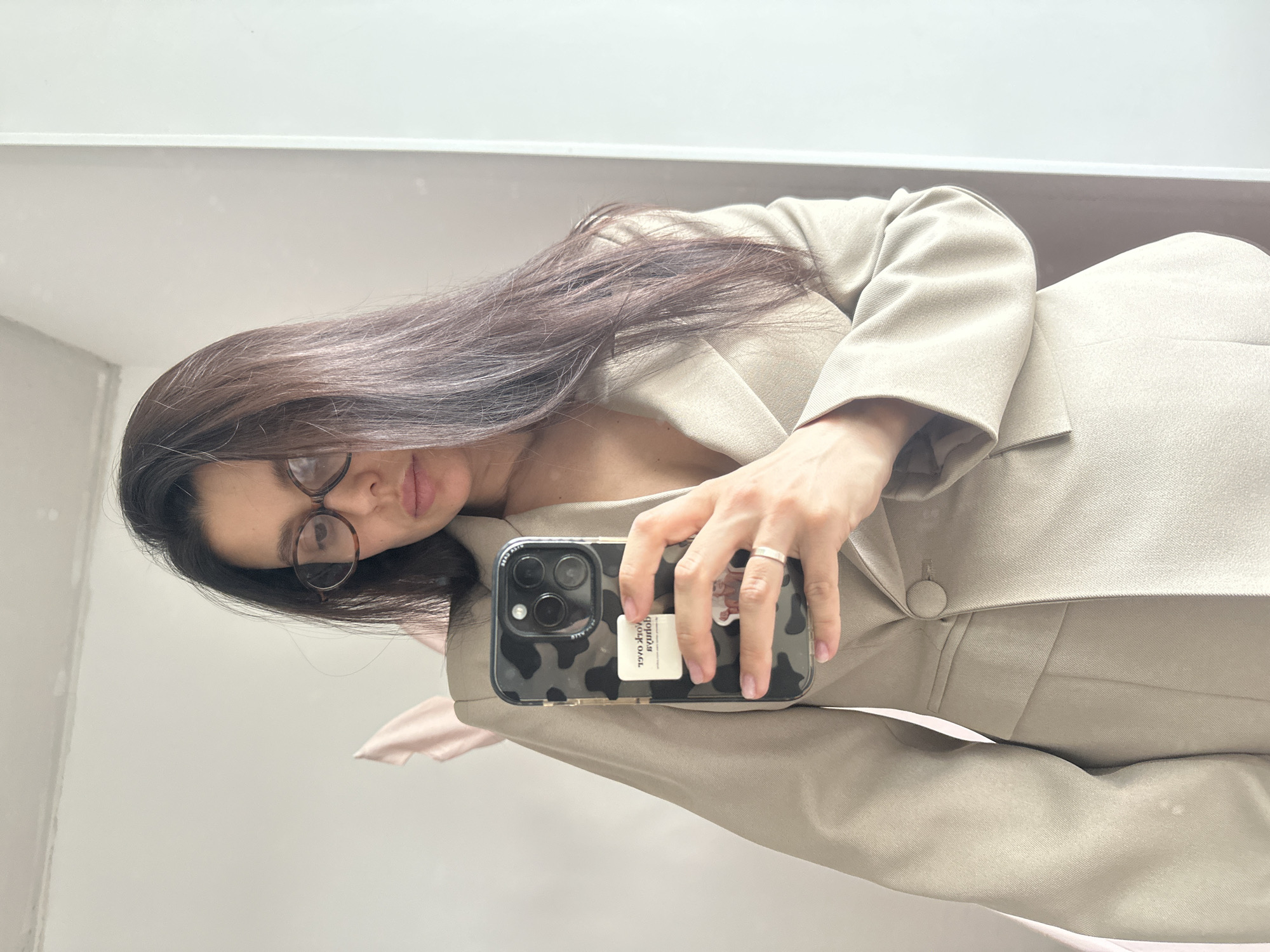 A woman is taking a selfie in front of a mirror. She is wearing glasses and a beige blazer. Her hair is long and brown. She is holding a black smartphone with a cow-print case. The mirror reflects a white wall. It is not possible to determine the location.