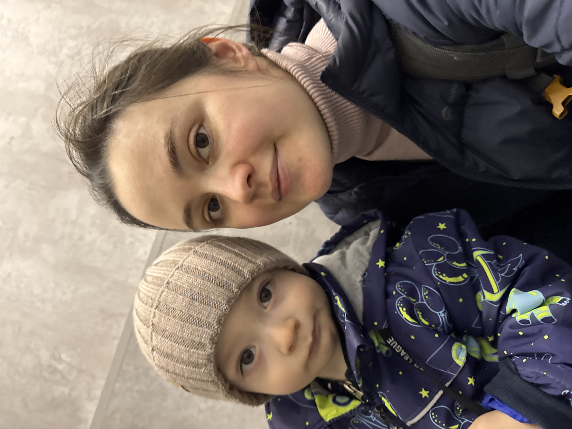 A woman and a toddler are posing for a selfie. The woman has brown hair pulled back, wearing a pink turtleneck and a navy puffy jacket. A portion of a backpack strap is visible behind her. The toddler is wearing a beige knitted hat and a navy blue jacket with a yellow print that includes stars and abstract shapes. They are likely inside, given the neutral-colored wall in the background. There is no specific information to suggest the location is Izhevsk, Russia.