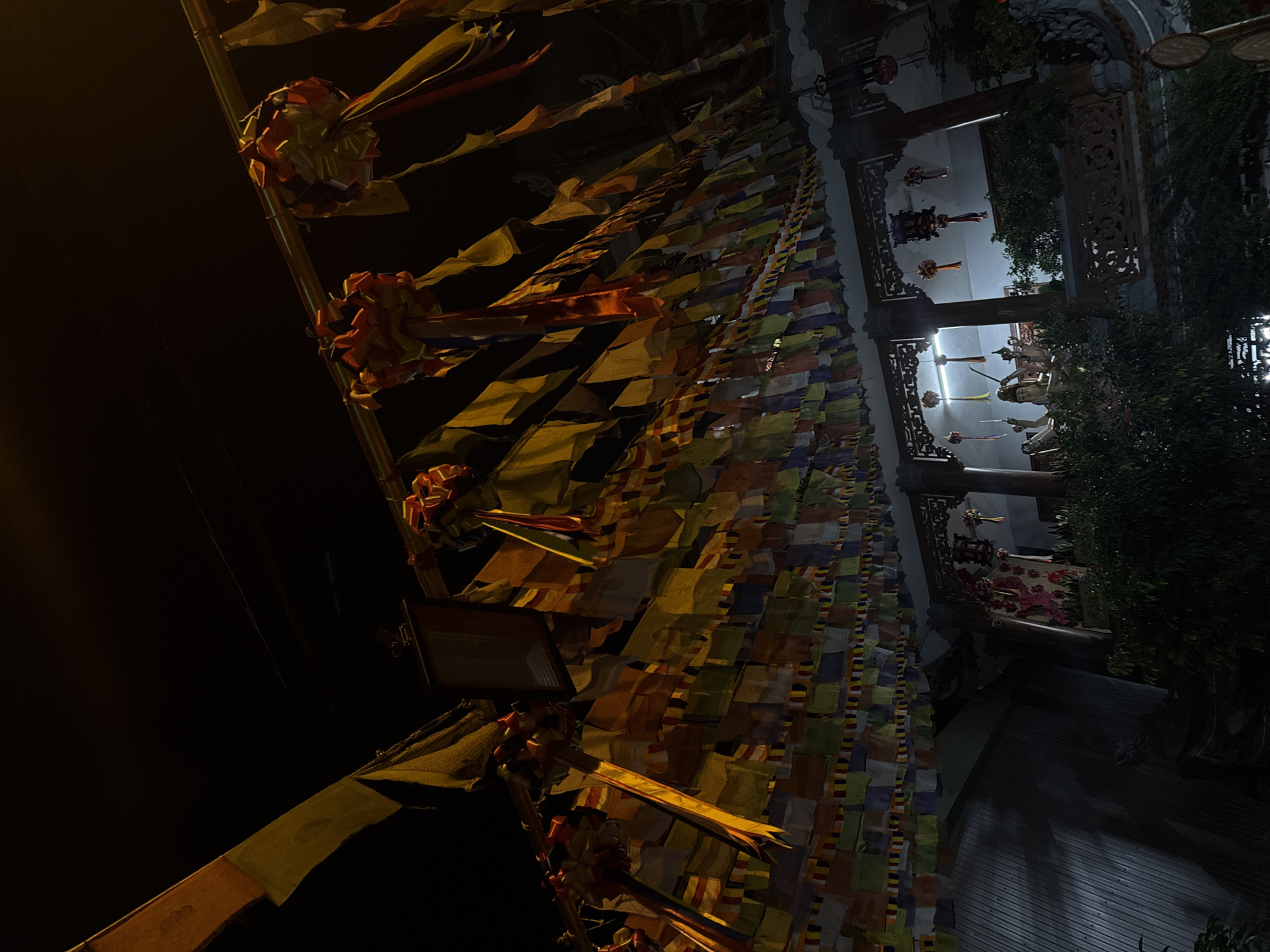 The image shows a nighttime scene in Đà Nẵng, Vietnam. Strings of prayer flags line the street in front of a temple or pagoda. The flags are illuminated by artificial light. Details of the temple's structure and design are visible, including wooden columns, decorative elements, and statues. Vegetation partially obscures the view.