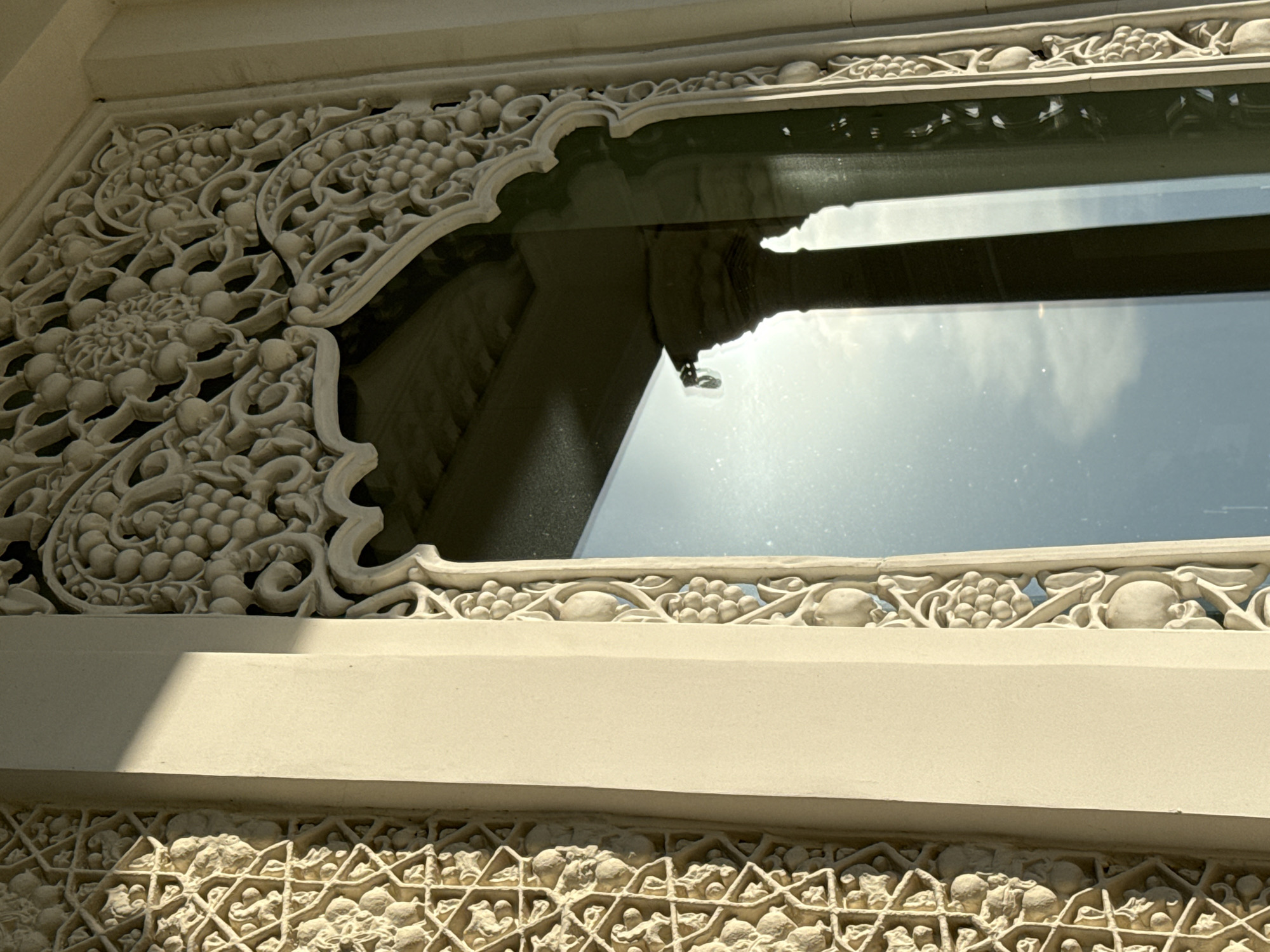 A detailed view captures a section of the intricate exterior architecture of the Cathedral of Christ the Savior in Moscow, Russia. The architectural detail includes a window framed with decorative, off-white or cream-colored stone carvings. The carvings display floral and vegetal motifs, including stylized grapes and leaves. The smooth, flat surfaces of the architecture suggest a recent cleaning. The window reflects the surrounding environment, showing the reflection of a building and the sky, indicating a bright, sunny day. The overall lighting is diffused, emphasizing the three-dimensional quality of the carvings.