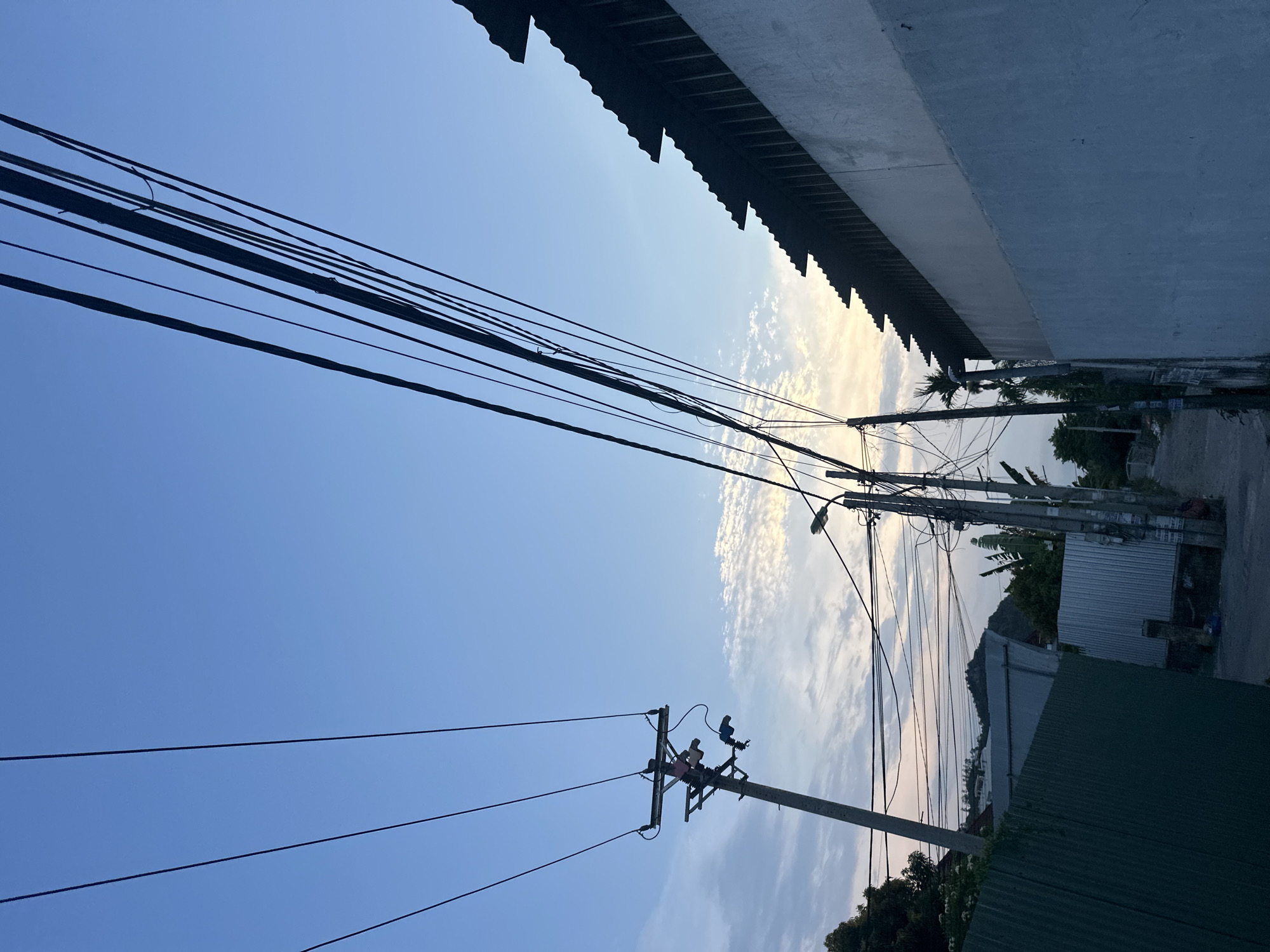 The image shows a street scene in Bac Nha Trang Ward, Vietnam. Power lines crisscross the sky, with a building overhang in the top right. Two power poles are visible, one closer to the bottom left of the frame. A street lamp is on a pole further down the street. Buildings and foliage line the street. The sky is blue with patches of cloud.