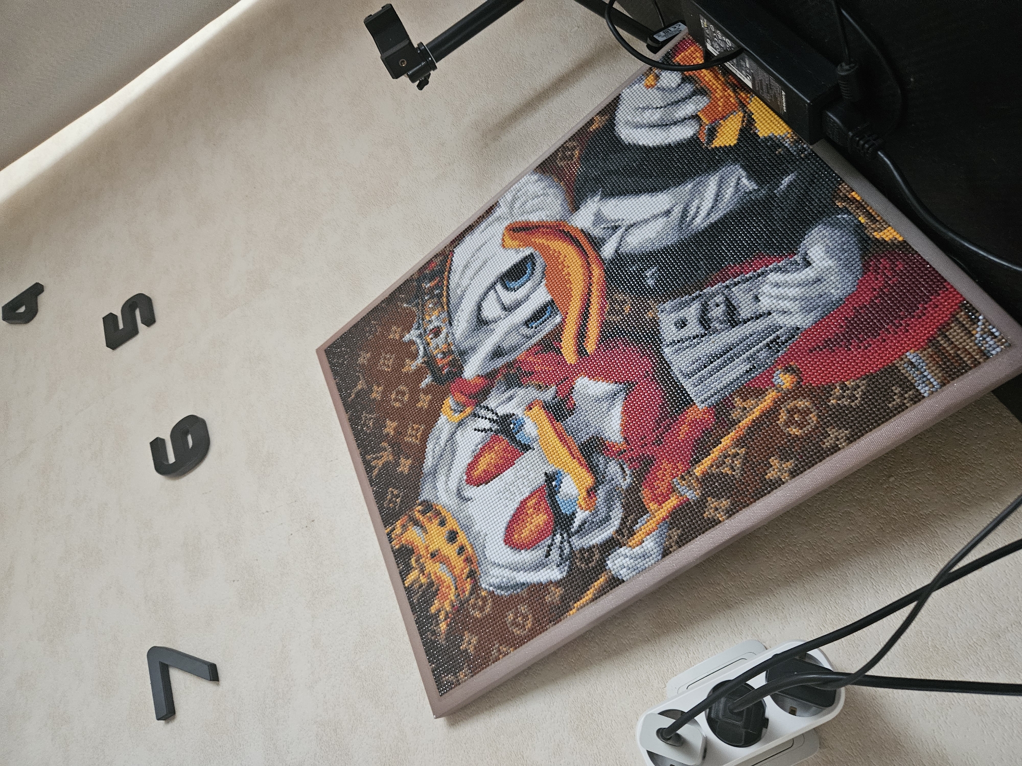 The image depicts a framed diamond painting featuring cartoon ducks wearing crowns, positioned on a textured wall. The diamond painting is leaning against the wall and has a light pink border. To the left of the painting, a white power strip with three outlets is plugged into the wall, with black electrical cords connected to it. Above and to the left of the painting, the numbers "7" and "6" are affixed to the wall. Further up and to the right, the numbers "4" and "5" are also visible on the wall. A black camera tripod with a camera attached is positioned to the right of the diamond painting, with black cords extending from it. The lighting in the scene suggests an indoor environment, with the wall having a light, speckled texture. The provided context indicates the location is Sosnogorsk, Russia, although the visual elements do not offer specific identifiers of this location.