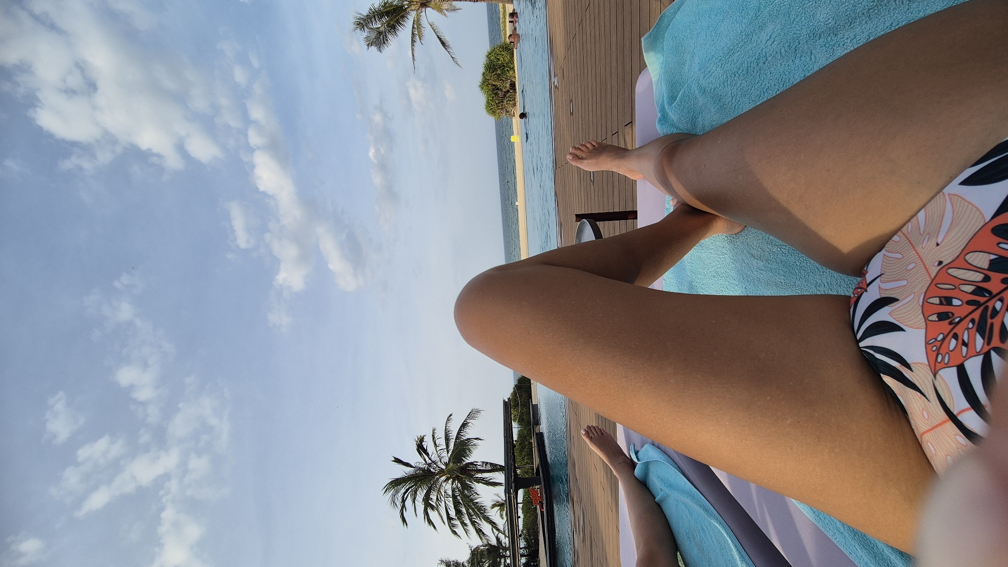 The image shows a first-person perspective of someone lying on a lounge chair by an infinity pool, with the ocean visible in the background. Palm trees frame the scene, and the sky is filled with scattered clouds. The person is wearing a bikini with a tropical leaf pattern. Another person's legs are visible on a lounge chair to the left. The poolside is made of wooden decking. There are several people in the pool in the distance. The location appears to be a resort in Hoi An, Vietnam.