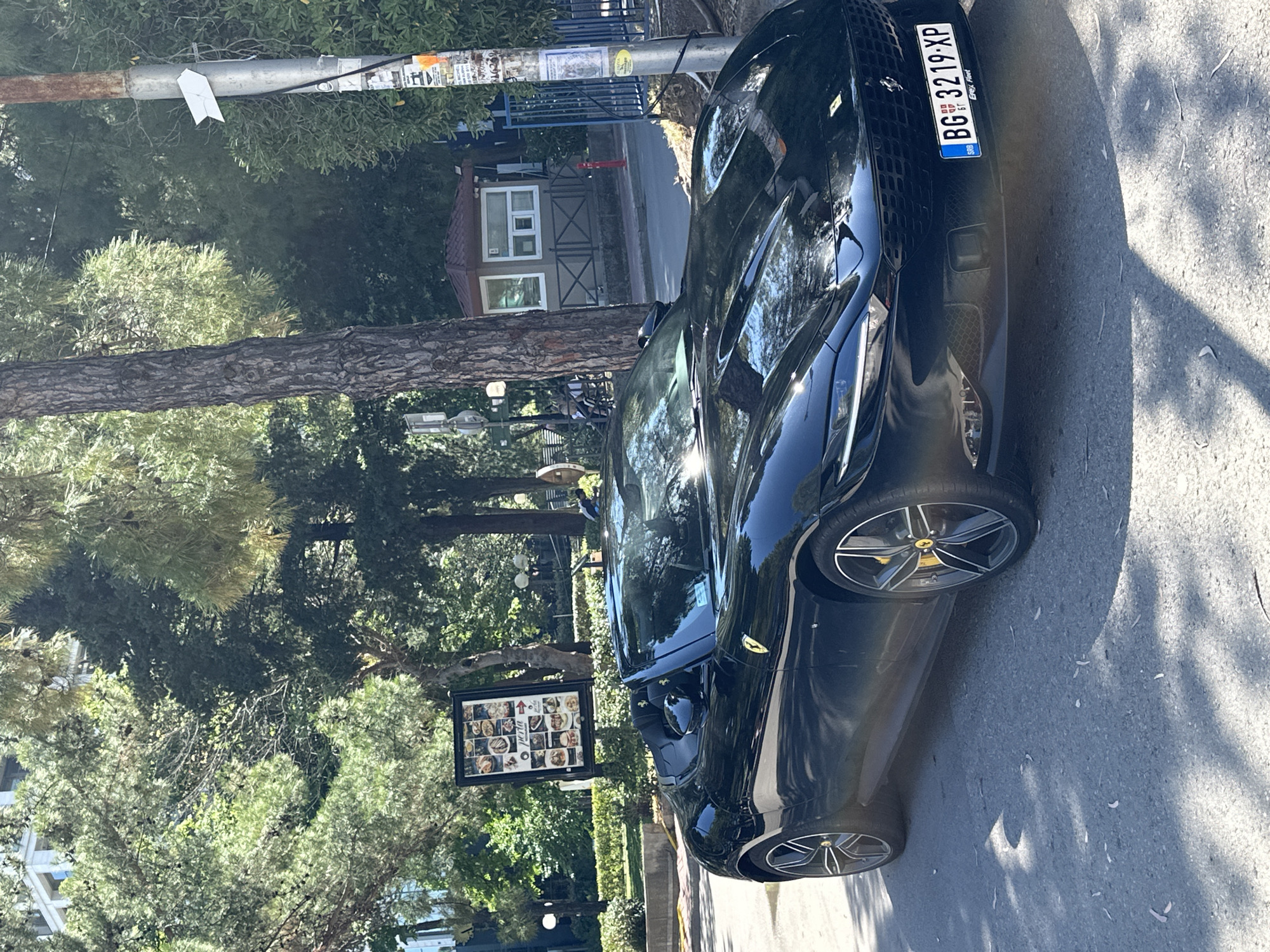 A black convertible Ferrari Roma is parked on a street in Budva, Montenegro. The car is facing forward and its front license plate reads "BG 3219-XP". The car is a convertible with the top down, revealing the black interior. In the background, there are trees and buildings. A menu board for a restaurant called "Perla" is visible to the left of the car. A lamppost with various stickers is to the right of the car. A small building that appears to be a security booth or kiosk is visible behind the car. The street is paved with asphalt, and there are shadows from the trees on the ground.