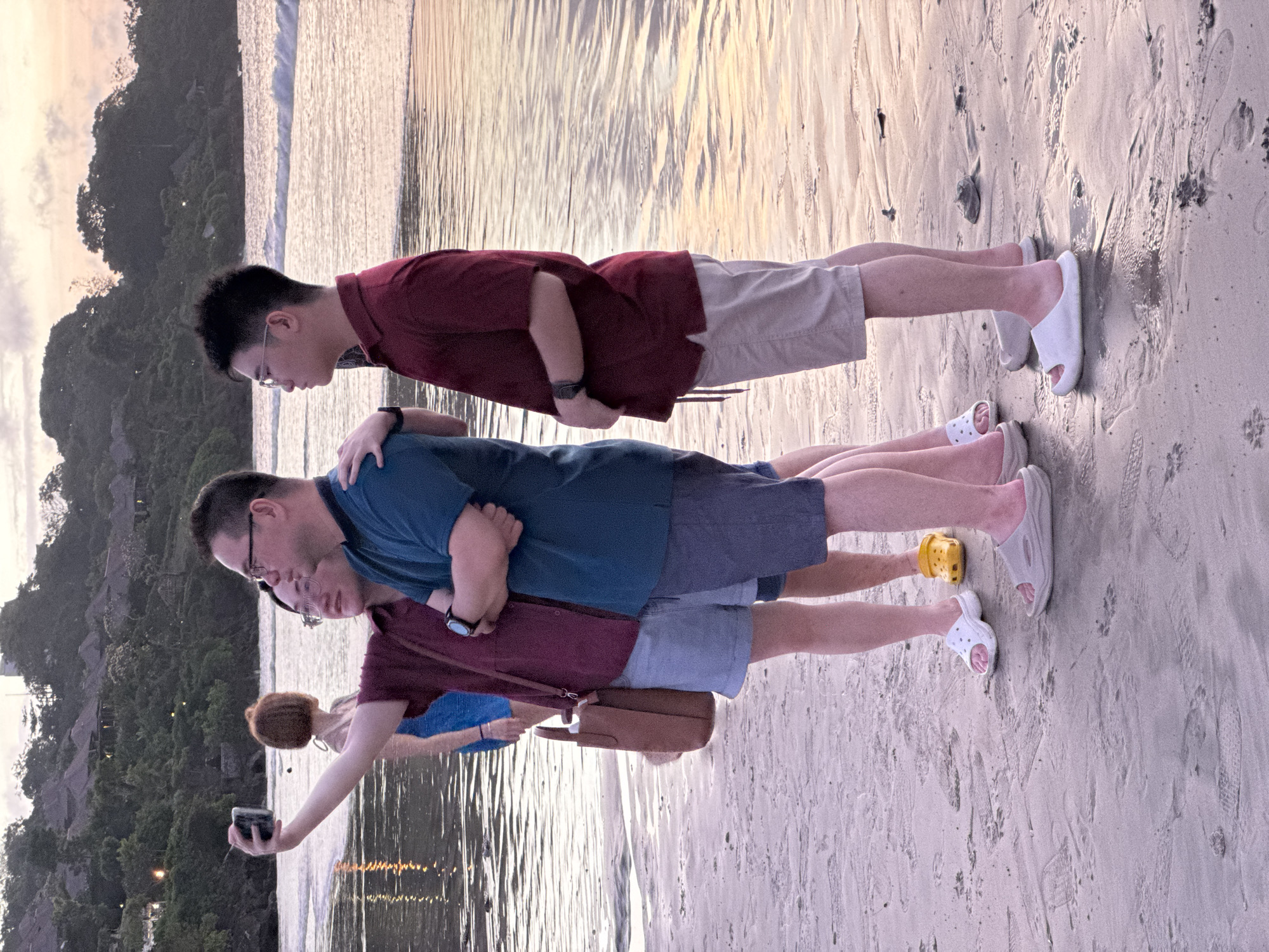 The image shows four people on a beach at dusk. The sky is overcast with soft light. Two men stand side-by-side, one with his arm around the other. A third person, possibly a woman with short, reddish-brown hair, is to their left, holding a smartphone to take a selfie. The fourth person is partially obscured behind the two standing men. They are all dressed in casual attire, with shorts and t-shirts. White sandals are visible on two of the individuals, and one person wears yellow slides. The ground is sandy with footprints. In the background, a hillside covered in lush green vegetation and some structures are visible. The sea is calm, with gentle waves lapping at the shore. The overall mood appears relaxed and social, possibly during a vacation.