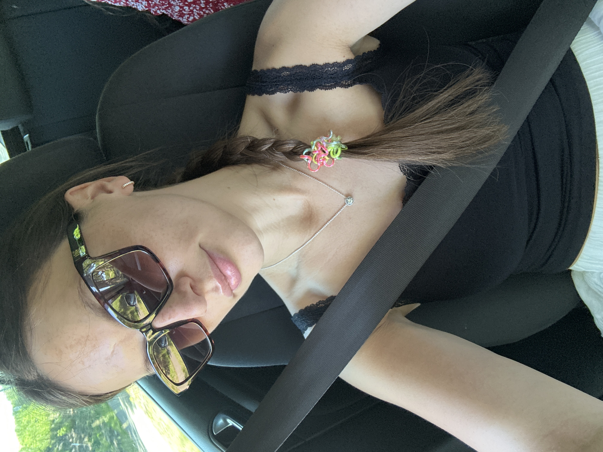 A selfie taken from inside a vehicle. The person is wearing a black top with lace trim, sunglasses, a delicate necklace, and has her hair braided with a colorful hair tie. She is wearing a seatbelt. The environment visible through the window appears to be outdoors with greenery. There is no information indicating the location is Stade, Germany.