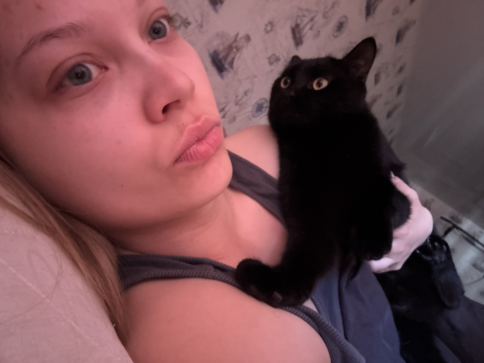 A woman is holding a black cat. The woman has light skin, blonde hair, and blue eyes. Her lips are pursed. She is wearing a blue tank top. The cat is looking to the right with yellow eyes. The background includes a patterned wall. The photo was taken indoors. The location is unknown; there is no information in the image to indicate it is in Chaikovsky, Russia.