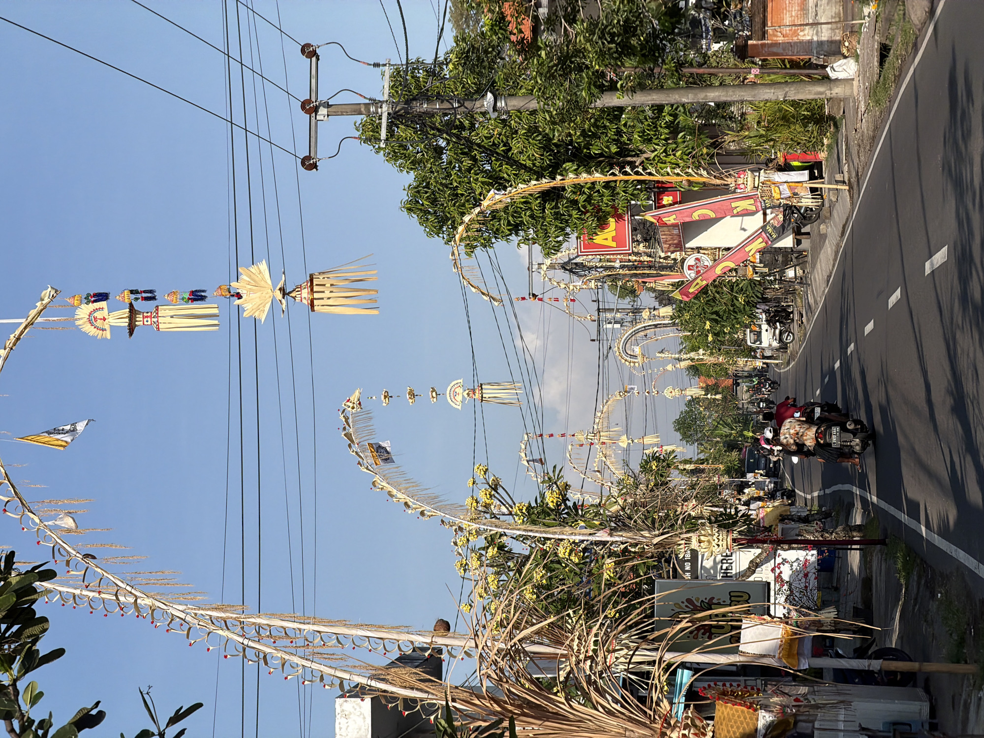 The image depicts a street scene in Mengwi, Indonesia, adorned with Balinese decorations for a religious ceremony. Ornate, palm frond arches and hanging ornaments, featuring woven patterns and colorful tassels, span the road. A clear blue sky serves as the backdrop, with power lines and utility poles visible overhead. Lush green trees and buildings line the street, suggesting a tropical setting. Several motorcycles are present, with one carrying two individuals moving along the asphalt road. The scene conveys a sense of cultural celebration and daily life in the region.