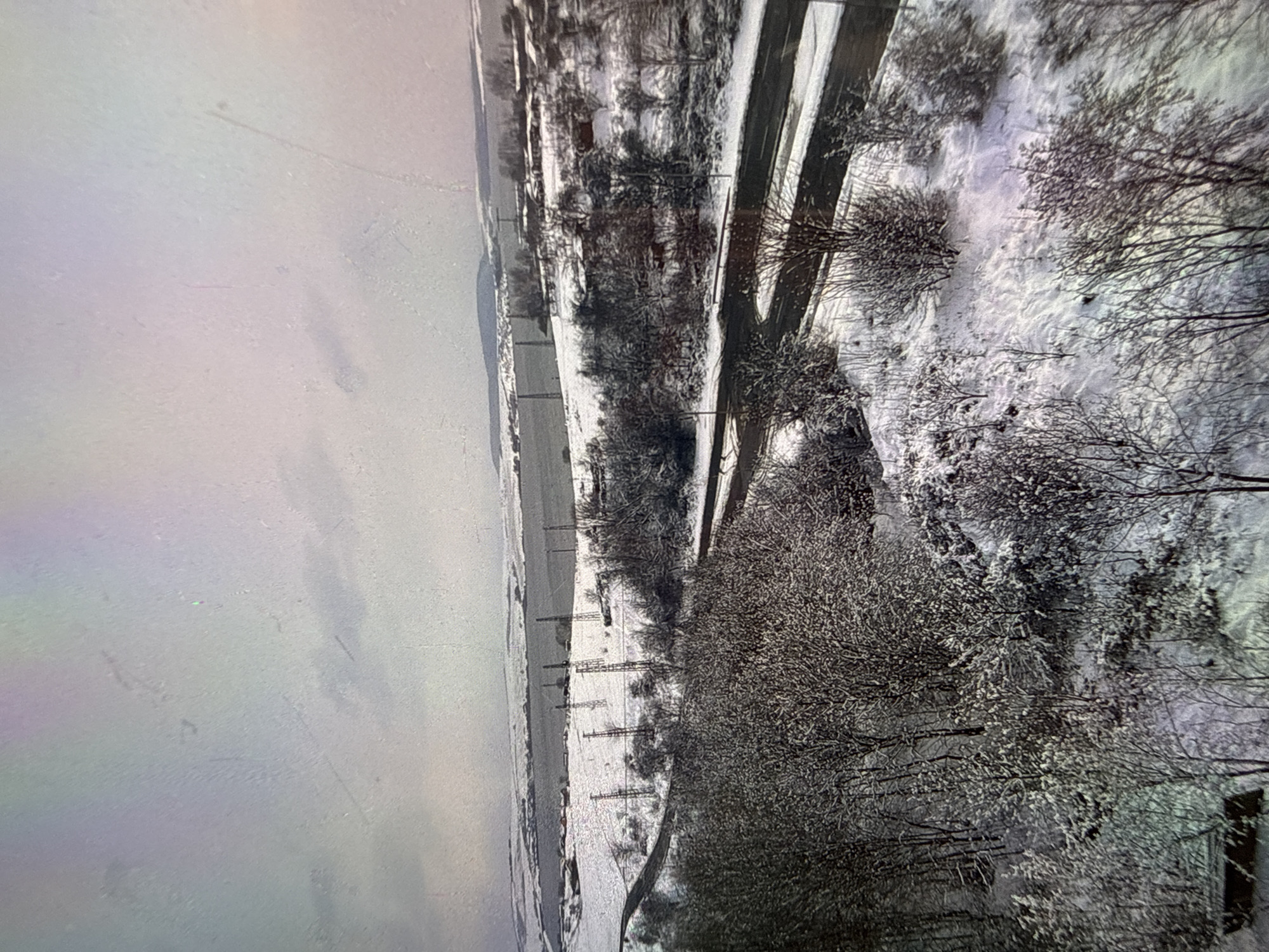 This is a media file depicting a winter landscape in Burgas, Bulgaria. The scene is dominated by snow-covered trees and fields, with a road running through the center of the frame. The overall impression is one of a cold, possibly overcast day, typical of winter weather.
The subjects are primarily natural elements: trees, snow, and a road. There are no visible people or animals. The road appears to be a relatively clear path through the snow, suggesting it might be a secondary road or one that is regularly maintained. In the background, there are hints of a flat landscape, possibly fields or open ground, also covered in snow. The trees are bare of leaves, a common sight in winter, and their branches are outlined by the white snow, creating a delicate, almost lace-like pattern against the darker ground and sky.
There is no specific activity or event taking place; the image captures a static, frozen moment in time. The time of day is difficult to ascertain precisely due to the overcast conditions, but the light suggests it is daytime, possibly late morning or early afternoon. The weather is clearly cold and snowy. The location cues, such as the mention of Burgas, Bulgaria, place the scene geographically.
Notable details include the texture of the snow on the branches, the linear perspective created by the road, and the vastness of the snow-covered terrain. The image evokes a sense of quiet and stillness, characteristic of a winter landscape. There is no visible text within the media file. The overall color palette is monochromatic, with shades of white, gray, and dark brown dominating the scene.
