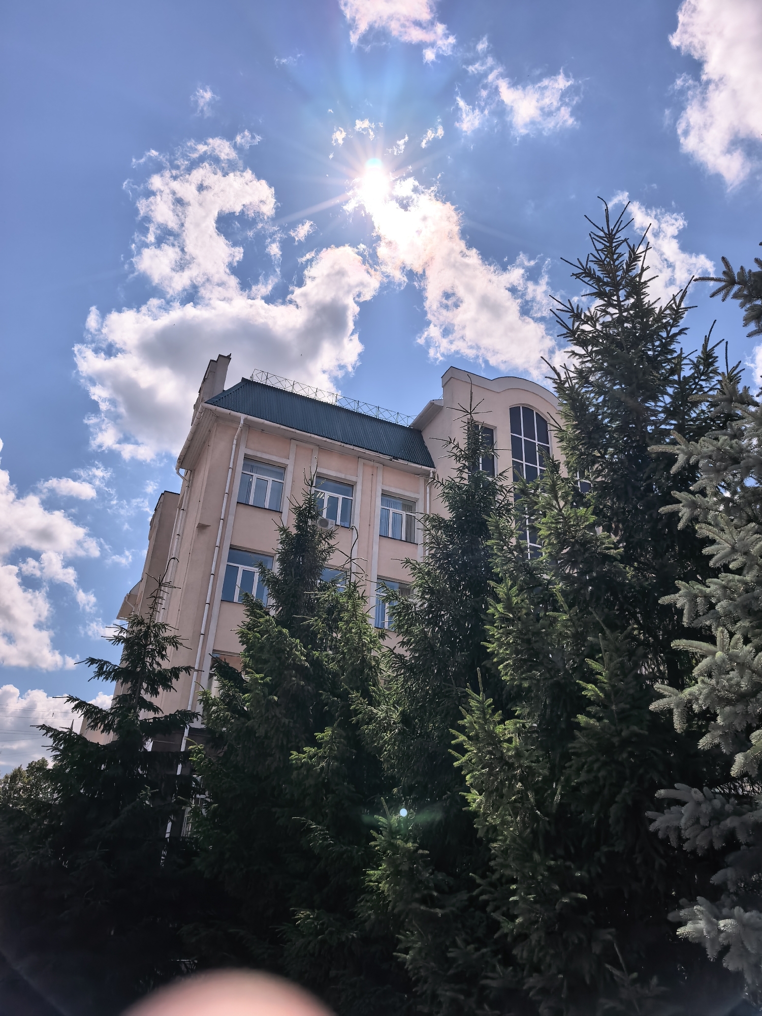 The image shows a multi-story building with a light pink facade and a dark green roof. The building has numerous windows with white frames. In front of the building, a dense row of evergreen trees, primarily spruce, obscures a significant portion of the lower levels. The trees vary in color from deep green to a lighter, bluish-green.
The scene is set outdoors, under a bright blue sky dotted with fluffy white clouds. The sun is prominently visible, shining intensely from the upper left quadrant of the image and creating a lens flare effect with visible rays of light. The presence of the sun and the bright sky suggest it is daytime, likely late morning or early afternoon, given the angle of the sun.
There are no people visible in the image. The activity, if any, is not apparent. The overall mood conveyed is one of a pleasant, sunny day in what appears to be an urban or suburban setting, possibly a park or a landscaped area adjacent to an institution or office building. The location context of Sterlitamak, Russia, provides a geographical reference but no specific visual cues within the image point to this exact location. No text is visible in the image.