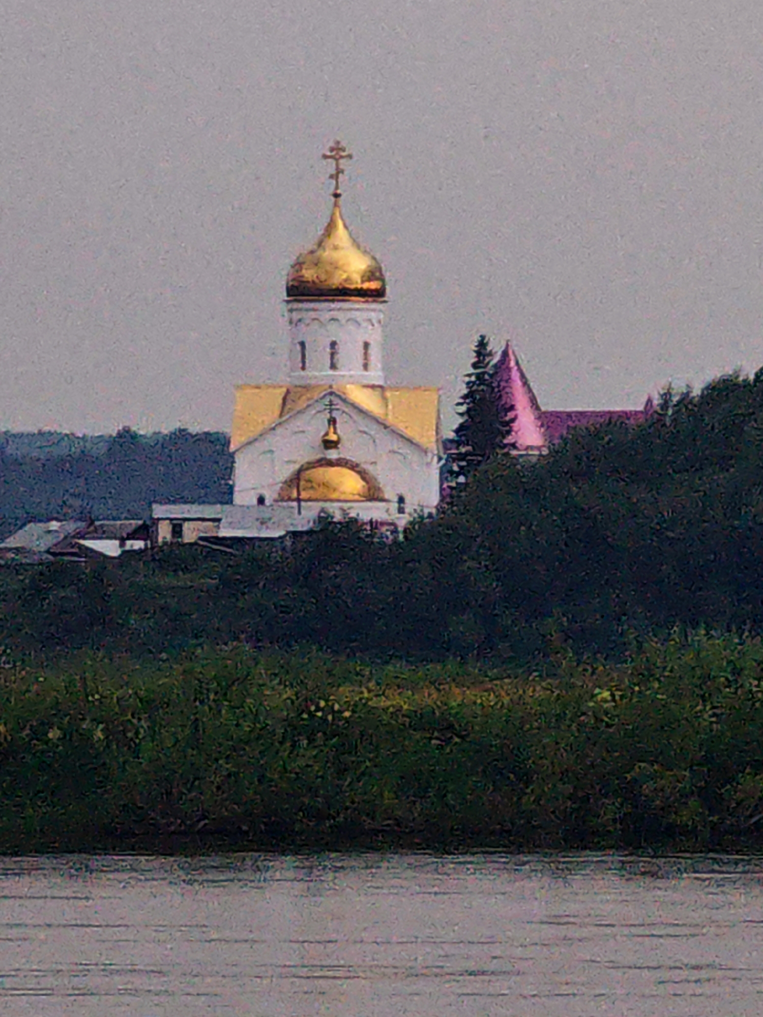 An Orthodox church with a golden dome and a gold cross on top stands prominently against a hazy, overcast sky. The white church has yellow trim and a smaller golden dome. To the right of the main church, a smaller building with a pink-hued roof is visible, partially obscured by dark green trees. In the foreground, a body of water with a reflective surface stretches across the bottom of the frame, bordered by lush green vegetation. The scene appears to be a landscape view of a settlement, possibly the village of Kazachinskoe, Russia, given the architectural style of the church. There are no people or discernible actions captured in the image.