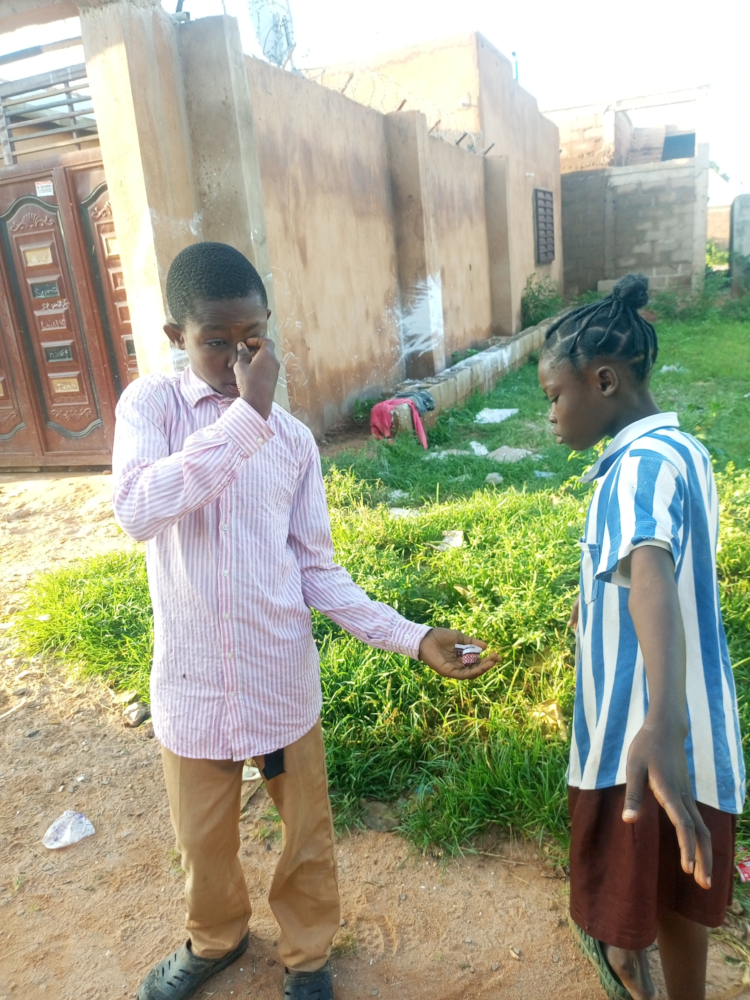 In Tempure, Nigeria, two young individuals are pictured outdoors during the daytime. The individual on the left, wearing a pink and white striped button-down shirt and tan trousers, is looking towards their right hand, which is held near their face, with their index finger touching their eye. They are also holding a small object in their outstretched left hand, presenting it to the person on the right. The individual on the right, dressed in a blue and white striped t-shirt and a brown skirt, is facing the person on the left and looking down at the object being offered. They have their hair styled in braids gathered into a bun on top of their head. The background consists of a dirt path, green grass, and a textured, light-colored building with some graffiti. A large wooden door with ornate carvings is visible to the left of the building. The scene appears to be in a residential or communal outdoor area.