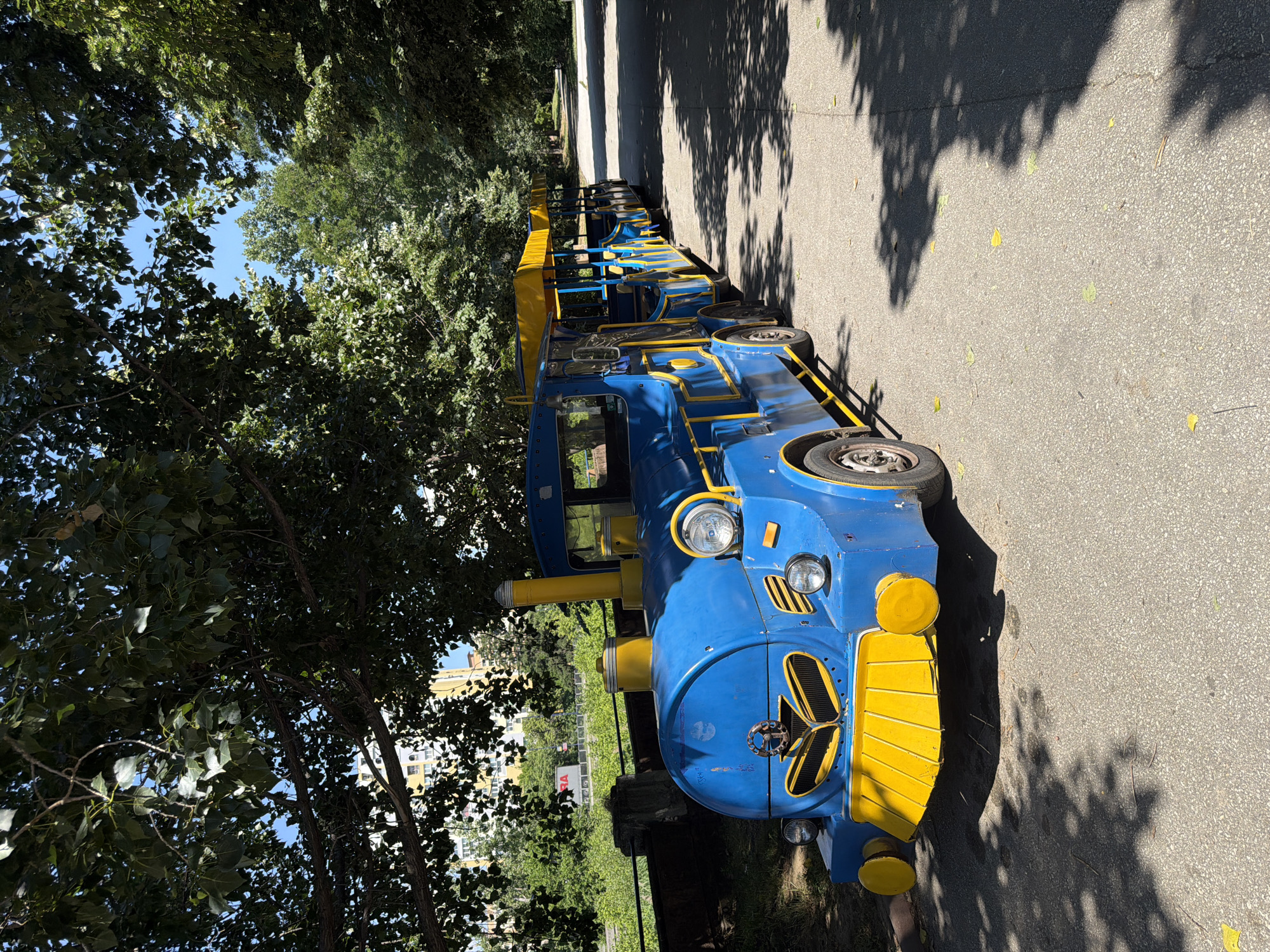 A blue and yellow amusement park train with multiple carriages is parked on an asphalt path in a park-like setting. The train is stationary, and no people are visible. The environment includes a paved walkway, large trees casting shadows, and lush green foliage. In the background, a multistory building and a red sign are partially visible behind the trees. The scene appears to be outdoors on a sunny day. The location is identified as Pazardzhik, Bulgaria.