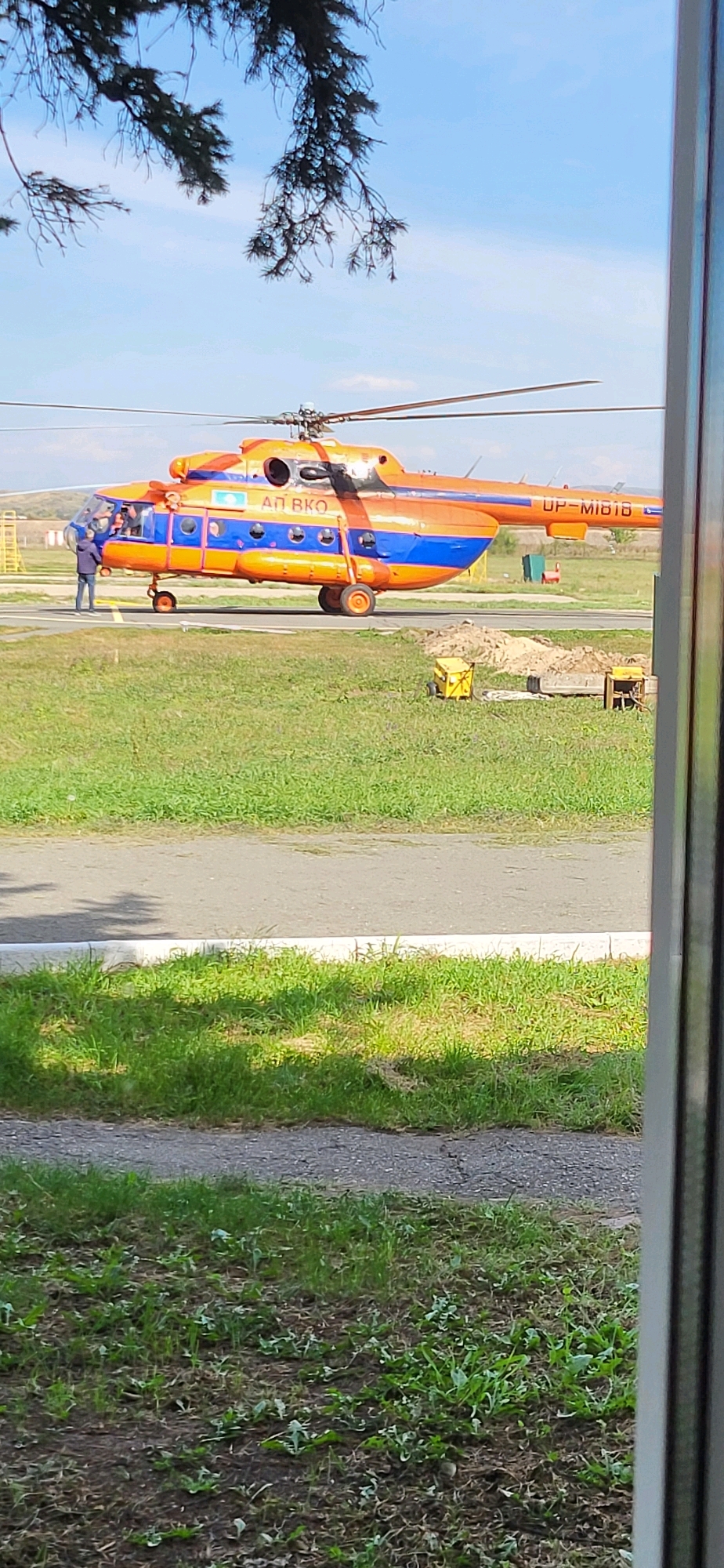 An orange and blue helicopter is parked on a tarmac with grass on either side. The helicopter has the registration "UP-MI818" on its tail. There is a person standing near the open door of the helicopter, wearing a blue jacket. In the background, there are some buildings and a yellow structure that looks like a ladder or a ramp. The sky is blue with some clouds. The scene appears to be an airport or airfield.