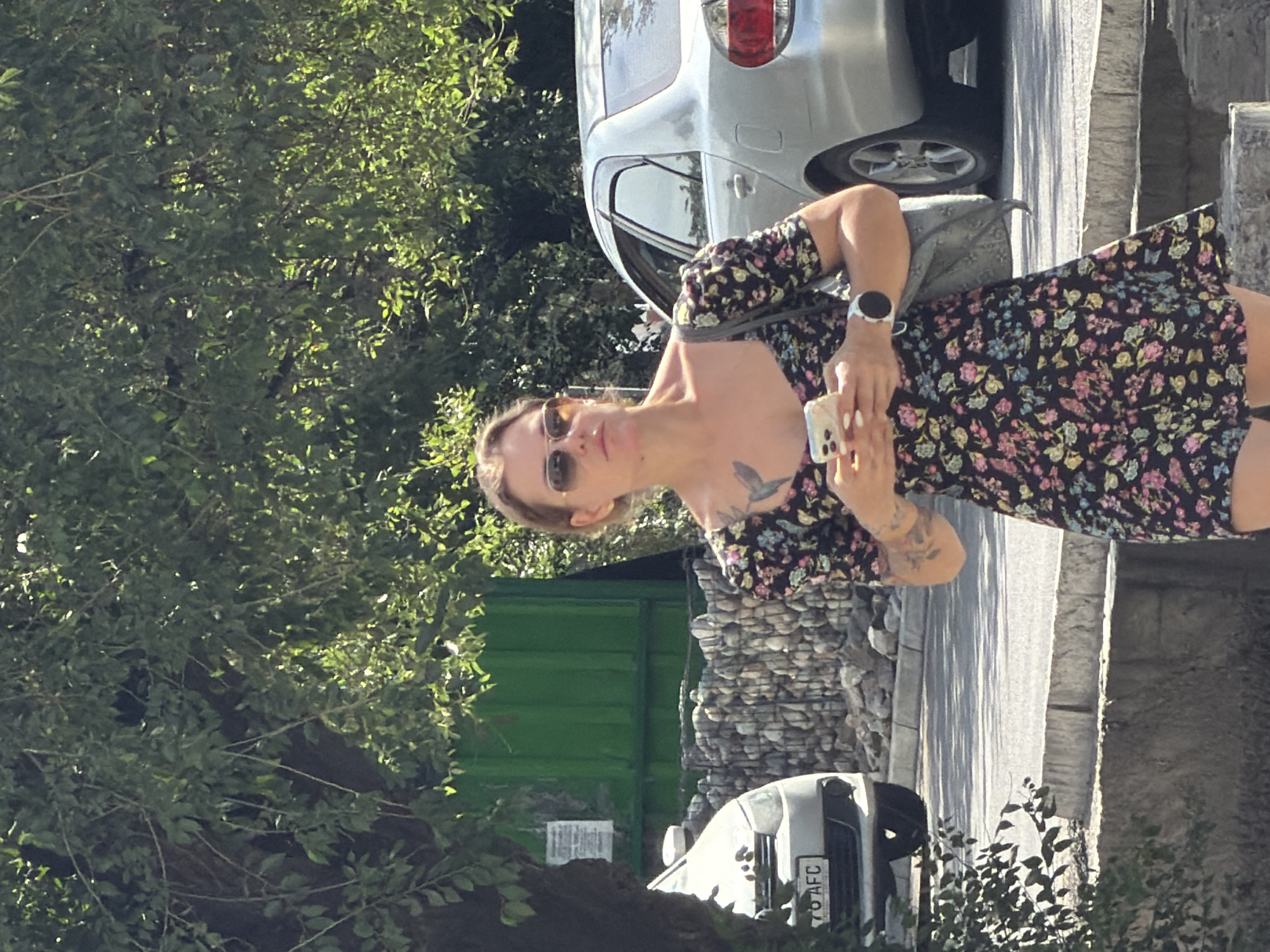 A woman with blonde hair pulled back and sunglasses on, wearing a dark floral print dress with puffed sleeves, is standing on a sidewalk. She is holding a white smartphone in her hands and looking down at it. She has tattoos on her left arm and shoulder. A grey car is parked behind her, and a silver car is to her left. A stone wall and a green building are visible in the background. The scene appears to be outdoors on a sunny day.