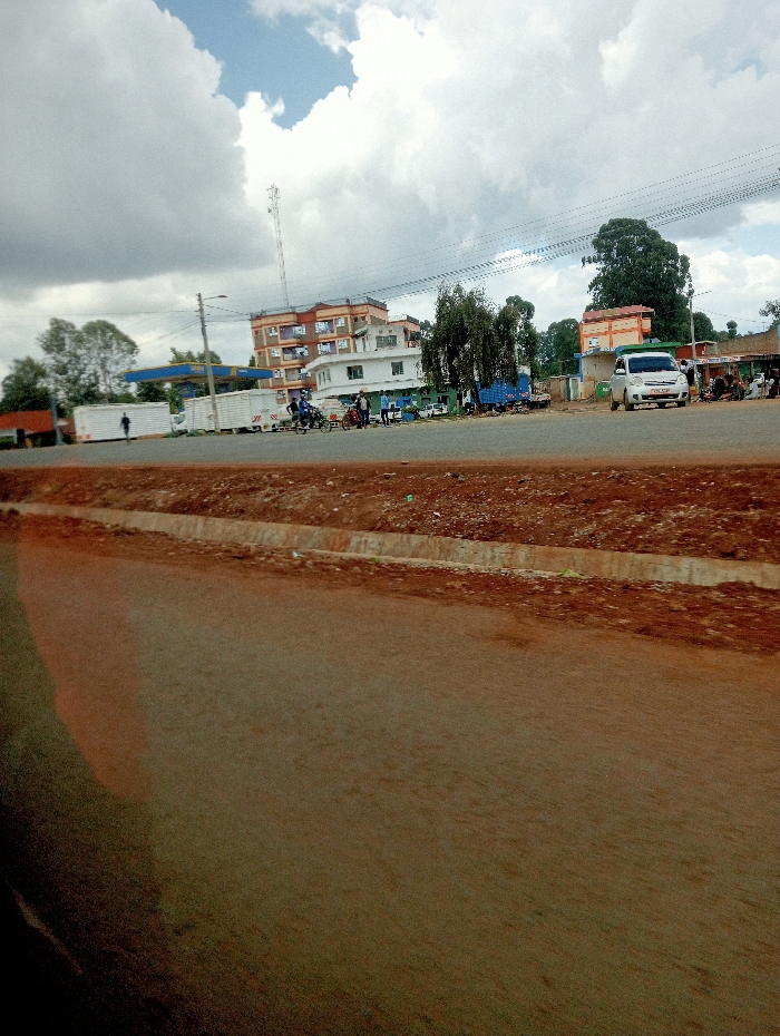 A street scene with several buildings and vehicles visible under a cloudy sky. The foreground shows a section of a road. There's a gas station and a tall communication tower. People are gathered near the roadside, and vehicles are parked along the street. Electrical wires stretch across the sky. The buildings vary in height, and some have multiple stories. The overall impression is of a busy, small-town environment.