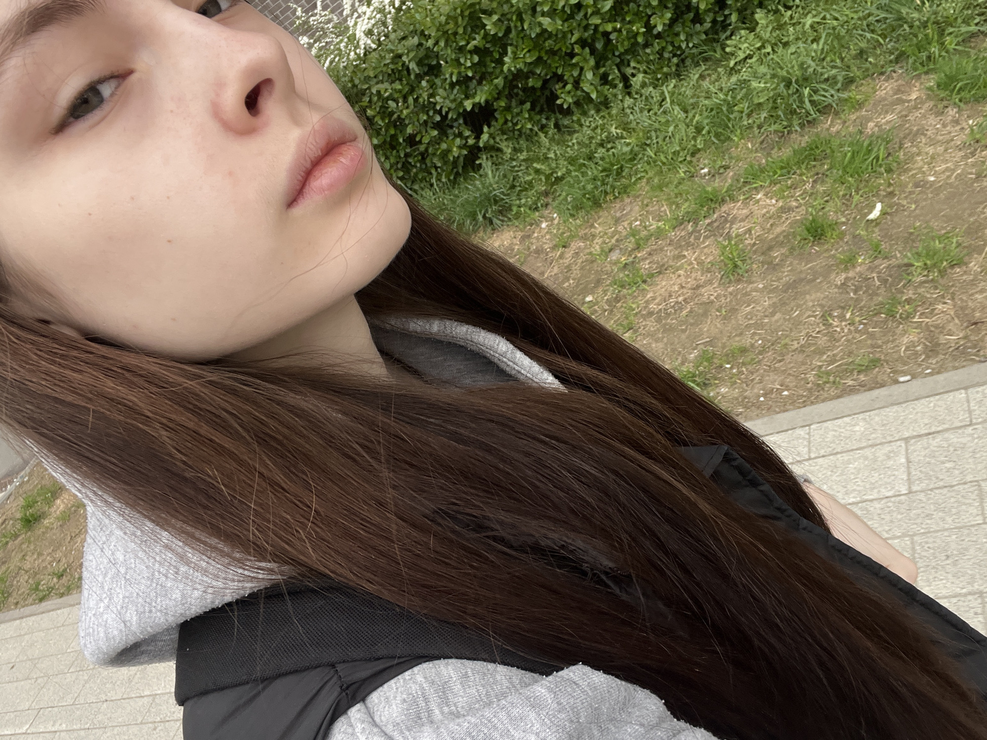 A selfie features a light-skinned individual with long, brown hair. The person wears a grey hoodie under a black vest. The background includes green foliage and a paved area, possibly a sidewalk or pathway. This likely takes place in a park or outdoor setting. The specific location within Saint Petersburg, Russia is impossible to determine solely from this image.