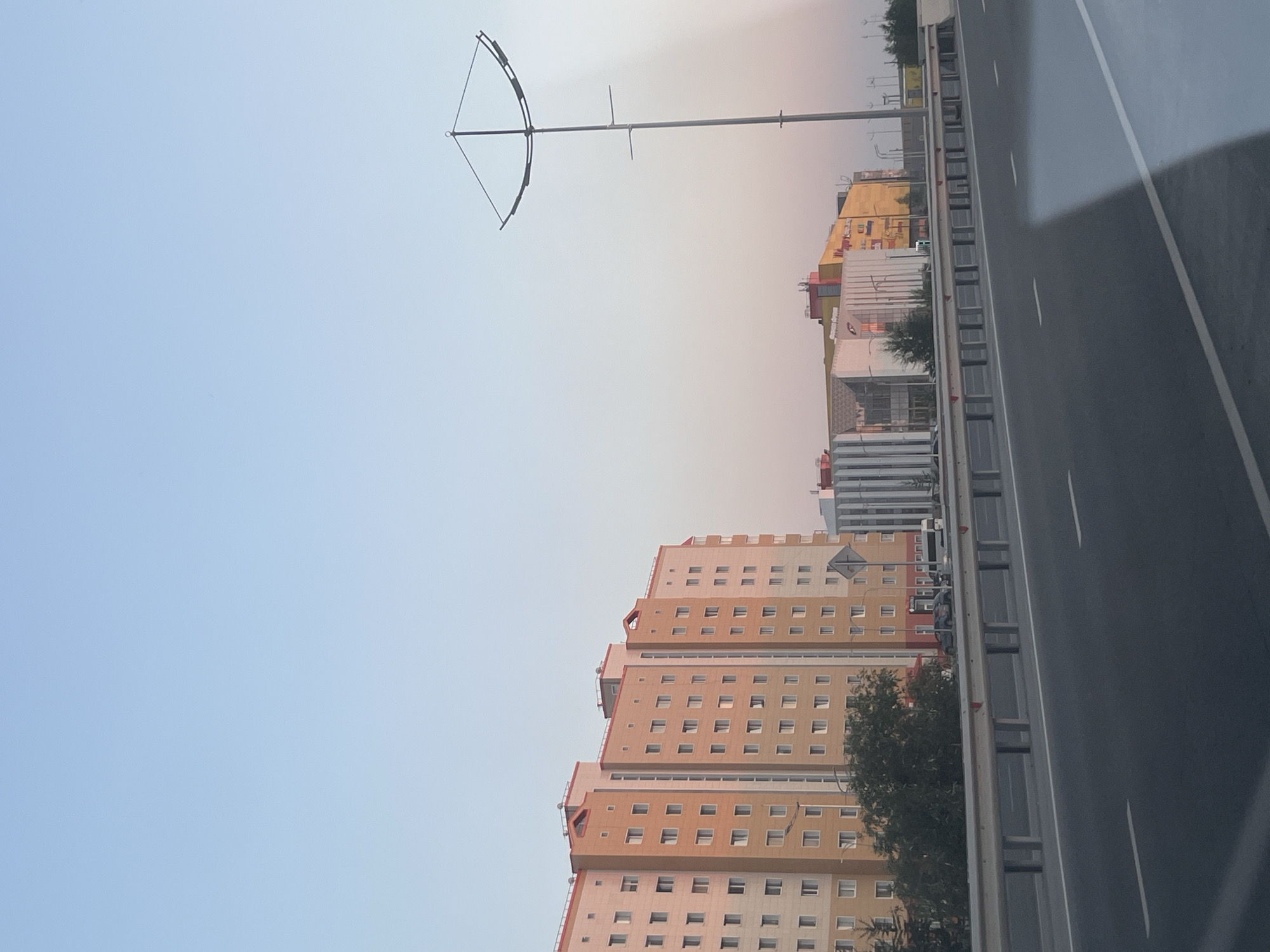 The image captures a section of a highway in Novy Urengoy, Russia, with a clear sky and several buildings in the background. On the left, a tall, multi-story apartment building with a predominantly orange and beige facade is visible. It features numerous windows arranged in a grid pattern. To the right of the apartment building, a series of modern commercial buildings with varied architectural styles and colors, including yellow, white, and pink, are present. One of these buildings appears to be a shopping center. A tall, distinctive street lamp with a curved top is positioned in the middle ground to the right. The highway itself is in the foreground, characterized by asphalt lanes marked with white lines and a metal guardrail separating it from the area beyond. A few trees and street signs are also discernible. No people or vehicles are clearly identifiable in action or interaction within the frame. The overall scene suggests a daytime view of an urban or suburban environment under a light blue sky.