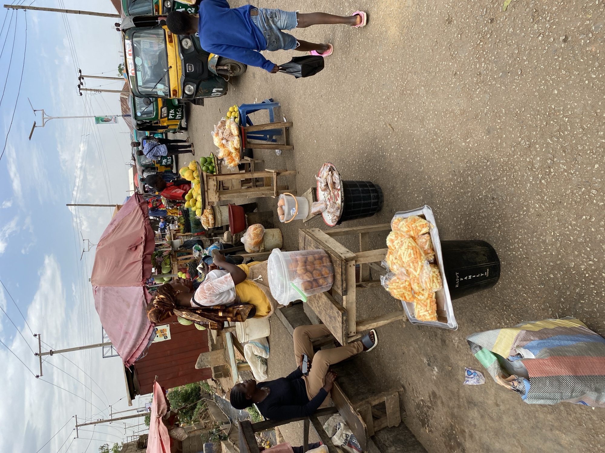 A street scene in Bukuru, Nigeria, shows market vendors and customers amidst various modes of transportation. The ground is paved. Vendors sell goods from makeshift wooden stands. A woman rests or sleeps near a stand with packaged snacks and a container of round, baked items. Another person sits nearby, appearing to be on their phone. A man in a blue shirt and shorts walks past, carrying a black bag. In the background, auto rickshaws are parked, and people mill about. Overhead, utility poles and lines are visible against a partly cloudy sky.