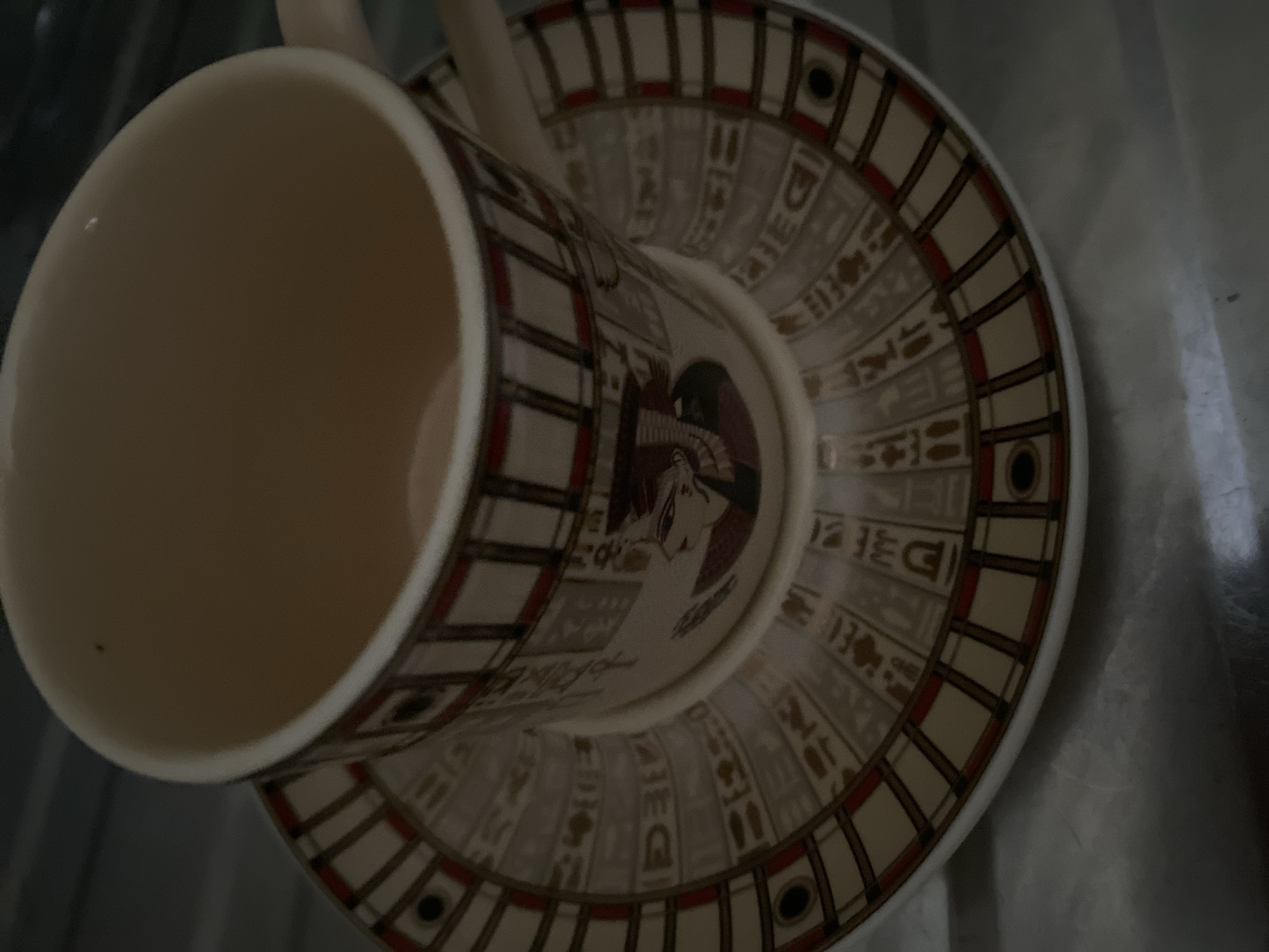An Egyptian-themed teacup and saucer are pictured. The cup is filled with a light brown liquid, and the saucer is decorated with hieroglyphics and a profile of an Egyptian pharaoh. The setting appears to be an indoor environment, possibly a kitchen or dining area, given the reflective surface beneath the saucer which could be a countertop or table. The lighting is dim, casting shadows and highlighting the glossy surfaces of the ceramic. There are no people or animals present in the image. The location in Sokhumi, Abkhazia is not evident from the visual information provided.