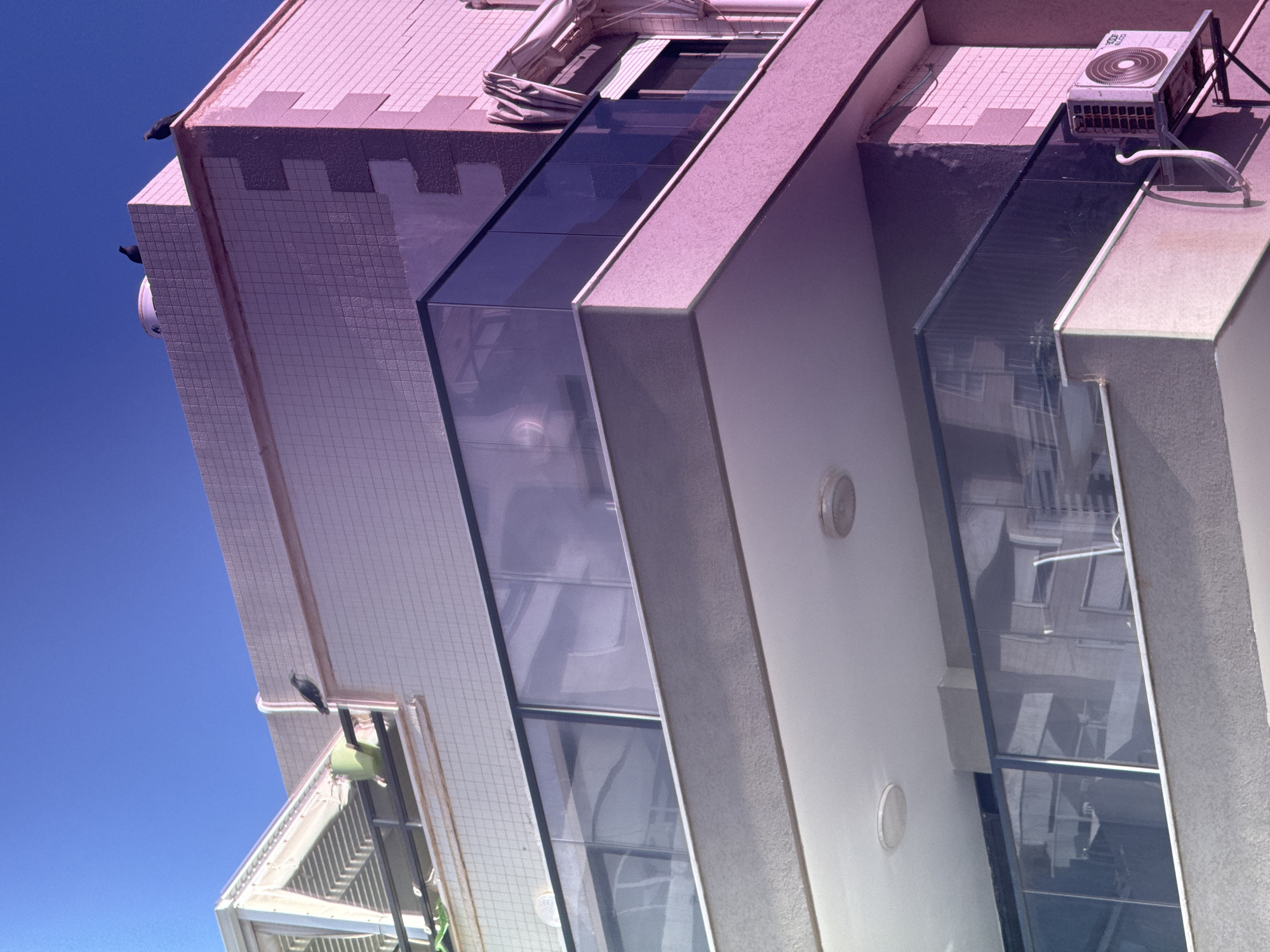 The image displays a rotated view of the exterior of a building, likely a residential complex in Bat Yam, Israel, against a clear, vibrant blue sky. The photograph, taken from a low angle and then rotated 90 degrees clockwise, presents a dynamic perspective of the architecture.
The building's facade is a mix of light-colored, possibly pale pink or beige, tiled sections and smooth grey/white concrete elements. Prominent vertical glass sections, acting as balconies or large windows, reflect parts of the building and surrounding structures, including other windows and architectural forms.
Under bright daylight conditions, evident from the strong shadows, the scene depicts a static moment. Two dark birds, resembling pigeons, are perched on the upper edge of the building's roofline, and another is visible near a lower window. An outdoor air conditioning condenser unit, grey with a circular fan, is mounted on a balcony or rooftop section. Indistinct white text is visible on the unit's casing. Other details include rolled-up fabric awnings above some glass areas, small circular light fixtures embedded in the walls, and some white piping near the AC unit.
The weather is clear and sunny, with no clouds in the sky. There is a subtle purple/pink color cast across the building, potentially from the lighting or image processing. No human activity or explicit emotions are discernible in the frame.