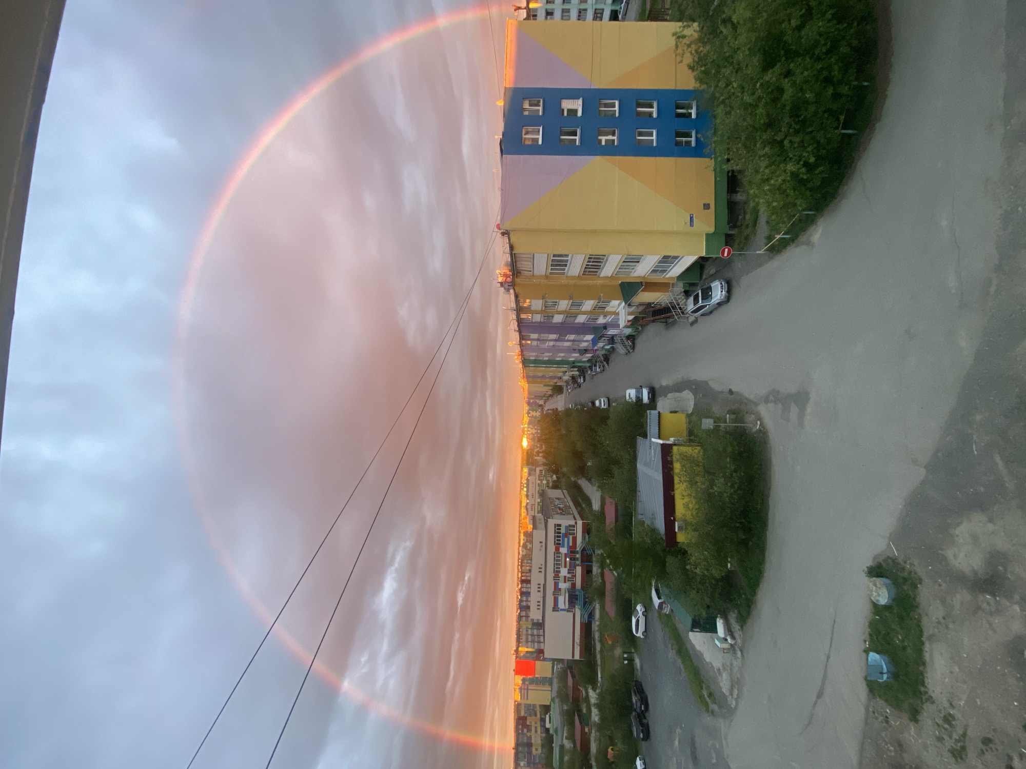 This image depicts an urban scene in Udachny, Russia, featuring a vibrant rainbow arcing across a cloudy sky at sunset. The setting is residential, with several multi-story apartment buildings dominating the landscape. The buildings display a variety of colorful facades, including yellow, blue, pink, and purple sections, with visible windows and balconies. A paved road runs alongside the buildings, with parked cars and some greenery interspersed.
The sun is low in the sky, casting a warm, orange glow on the clouds and the tops of the buildings, indicating it is either sunrise or sunset. The weather appears to be a mix of clouds and clearing skies, with the rainbow suggesting recent or ongoing precipitation. There are power lines running diagonally across the frame, adding to the urban feel. The overall mood is somewhat serene, with the natural phenomenon of the rainbow providing a striking contrast to the man-made structures. No people are clearly visible in the image. The location context of Udachny, Russia, suggests a northern climate, which could make such atmospheric phenomena particularly striking.