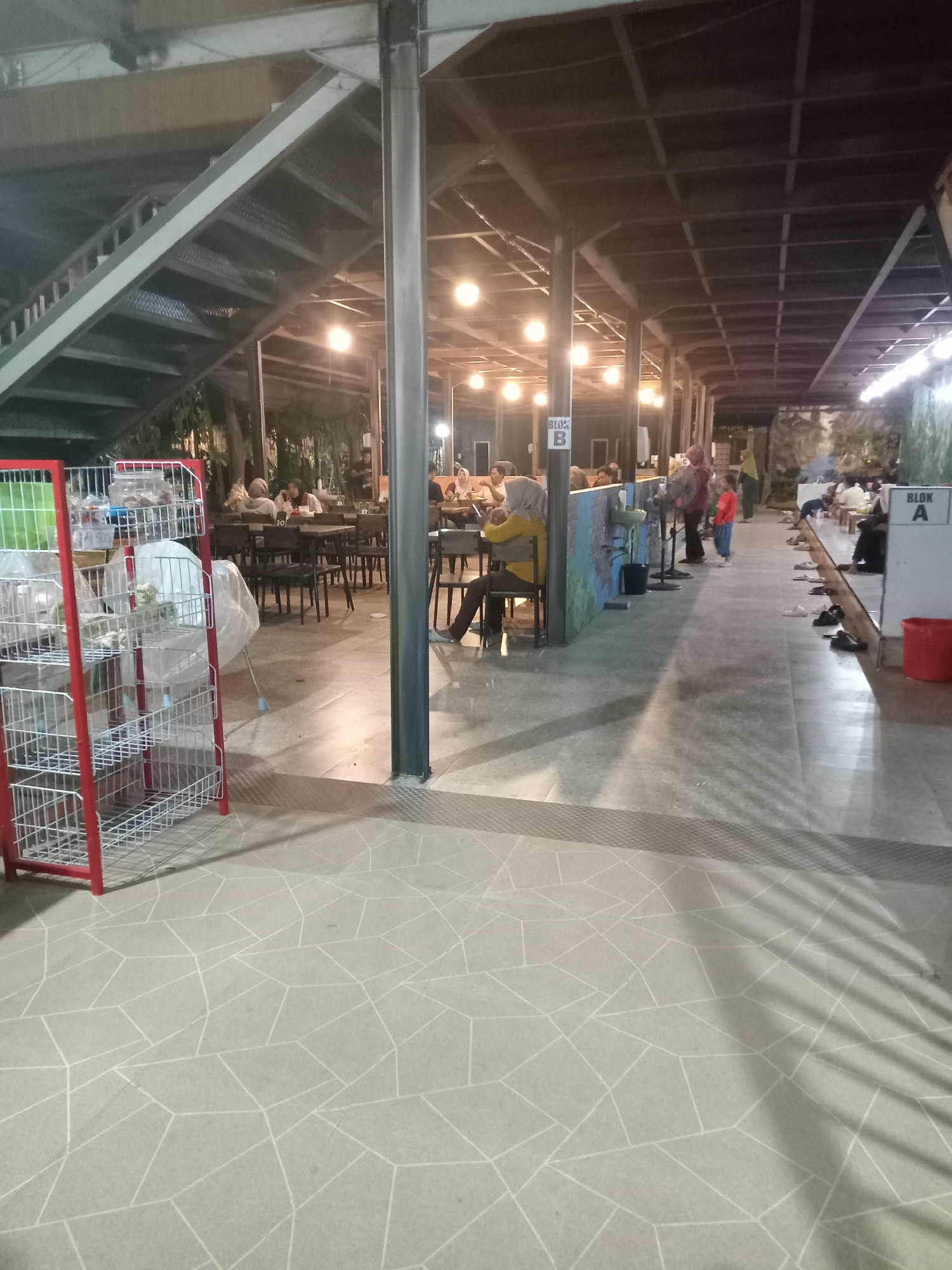 The image depicts an outdoor dining area in Sukomoro, Indonesia. Patrons are seated at tables in the background. A metal staircase is visible overhead on the left. A metal shelving unit with items covered in plastic wrap is in the foreground. Some individuals are queuing near a washing station labeled "Blok B". Further down the passageway, an area labeled "Blok A" contains seated people with shoes removed. The floor has a geometric tile pattern. The area is lit by overhead lighting.