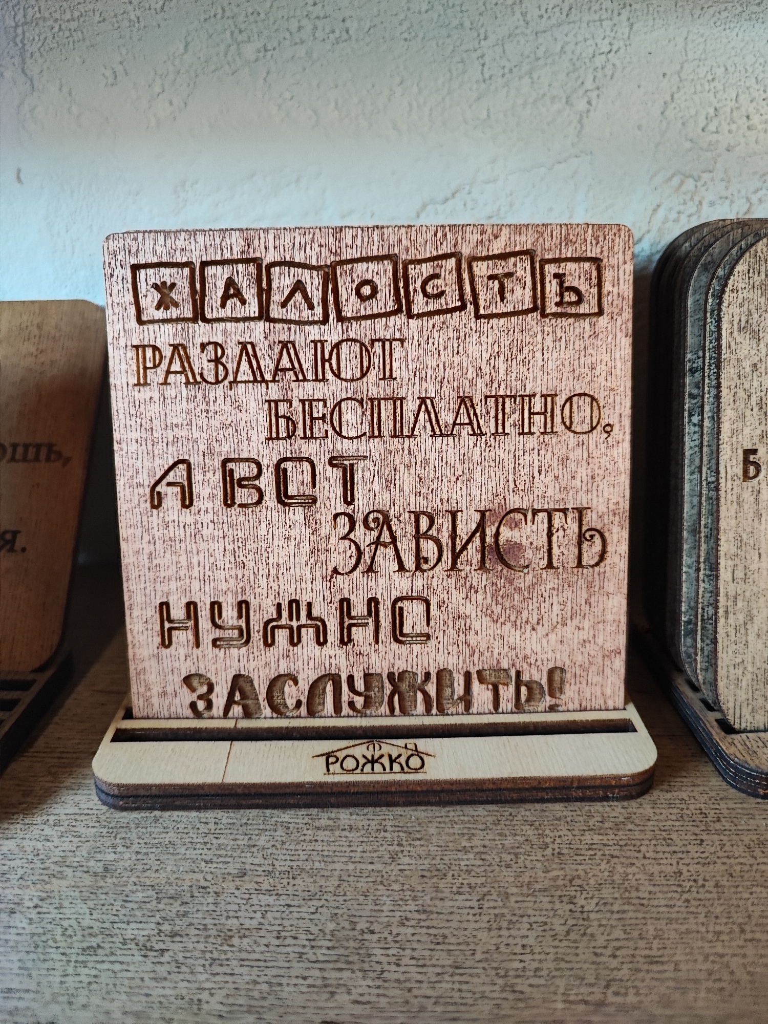 The image shows a wooden plaque with carved Russian text, displayed on a wooden shelf. The plaque is the central focus, with other wooden objects to its left and right. The text on the plaque reads, "ХАЛОСТЬ РАЗДАЮТ БЕСПЛАТНО, А ВОТ ЗАВИСТЬ НУЖНО ЗАСЛУЖИТЬ!" which translates to "Compliments are given for free, but envy must be earned!". Below the main text, a logo with the letters "РОЖКО" is visible. The plaque is supported by a wooden stand, also carved with the "РОЖКО" logo. The background appears to be a textured wall. The setting is an indoor environment, likely a shop or a display area. There are no people visible in the image. The text and the logo suggest that this is a decorative or humorous item. Pyatigorsk, Russia is a city known for its mineral springs and spa resorts, and it is plausible that such a souvenir or decorative item could be found there.
