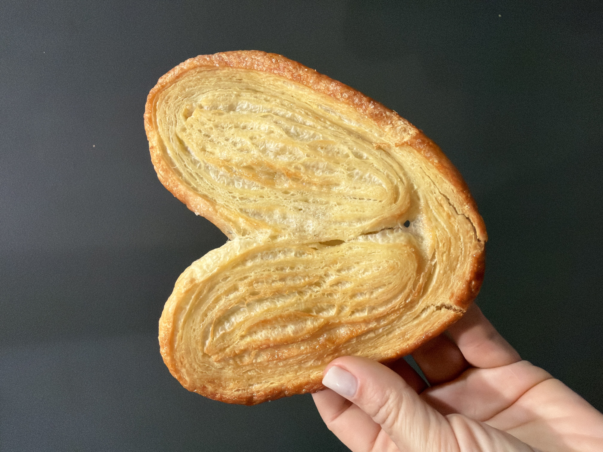 A single palmerita, also known as a palmier or elephant ear pastry, is held in a hand with a fingernail showing white polish, against a dark grey background. The pastry is golden-brown with a layered texture, indicating it is made of puff pastry. The location is not discernible from the image. There are no visible people besides the hand holding the object.