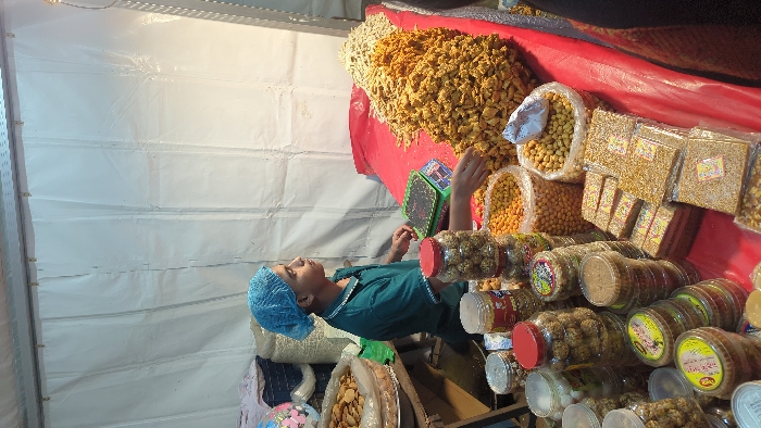 The image displays an indoor market stall, rotated 90 degrees counter-clockwise from its correct orientation. If rotated correctly, the view would be of a vendor standing behind a display counter laden with various snacks.
In the center of the image, a person, likely a vendor, is visible. They are wearing a dark teal or green short-sleeved top and a light blue hair net. Their hands are resting on the counter surface, near a dark green and black weighing scale. The person is looking upwards and slightly to their left, suggesting they might be observing something or someone outside the frame, or looking at a customer.
The stall itself is a vibrant display of food items. A large amount of what appears to be fried, savory snacks (yellowish-brown, irregularly shaped pieces) is piled high on a red cloth that covers the display counter. Next to these large piles are numerous clear plastic jars and bags filled with different dry snacks. Some clear plastic bags hold light-colored, spherical nuts, likely peanuts. Other clear plastic packets contain flat, rectangular blocks of what resembles sesame brittle or a similar confection, with visible seeds and possibly syrup.
Further along the counter, many clear plastic jars with red or white lids are neatly arranged. These jars contain a variety of items, including round, ball-shaped snacks (possibly sweets or fried dough balls) and granular mixtures. Several jars feature colorful labels, some predominantly yellow or green, with text in an Indic script (difficult to decipher specifically without higher resolution, but visible as local language). On one jar label near the bottom right, the weight "250g" is visible.
The background consists of plain white walls, which appear to be made of temporary material like tarpaulin or plastic sheeting, fastened to a frame, suggesting the setting is a temporary market, fair, or exhibition tent. The lighting appears artificial, illuminating the stall and its products. The overall impression is that of a bustling food stall ready for business, with a focus on a wide selection of packaged and bulk dry snacks.