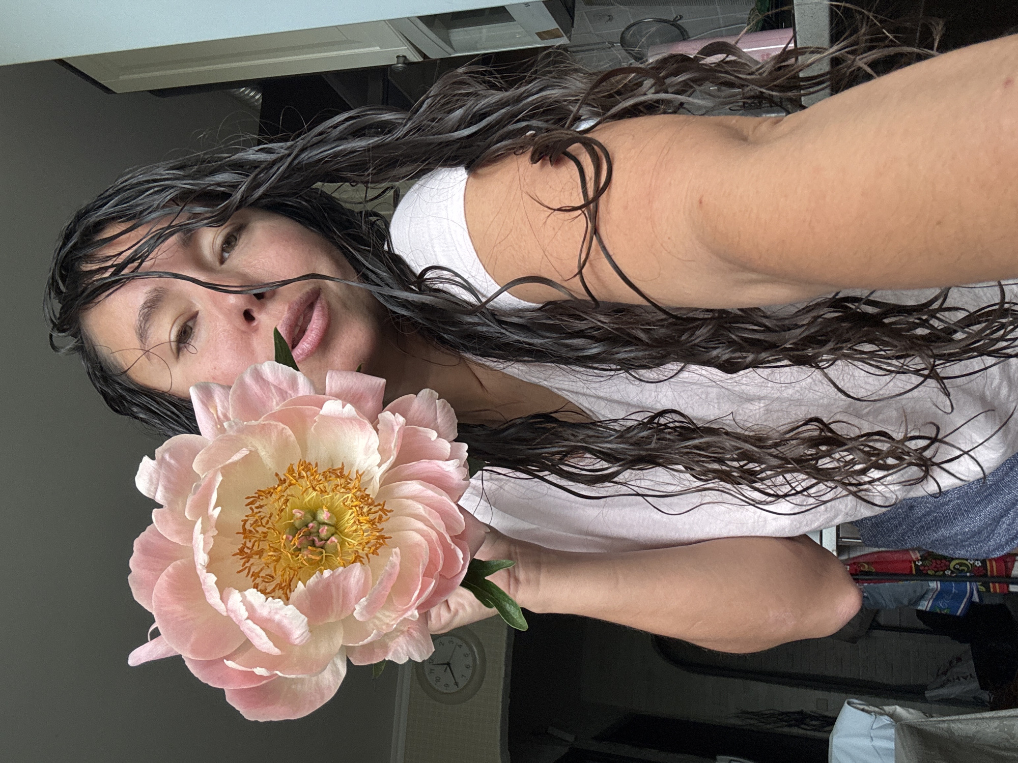 A medium shot captures a woman with wet, dark hair holding a large, light pink peony flower. She is positioned in the foreground, her face turned slightly towards the viewer, with her mouth slightly open. Her wet hair frames her face and cascades down her shoulders and arms. The woman is wearing a light-colored t-shirt. The background is indistinct but appears to be an indoor setting, possibly a kitchen, with a wall clock visible to the left. The lighting suggests an overcast day. The location is the city of Kazan, Russia.