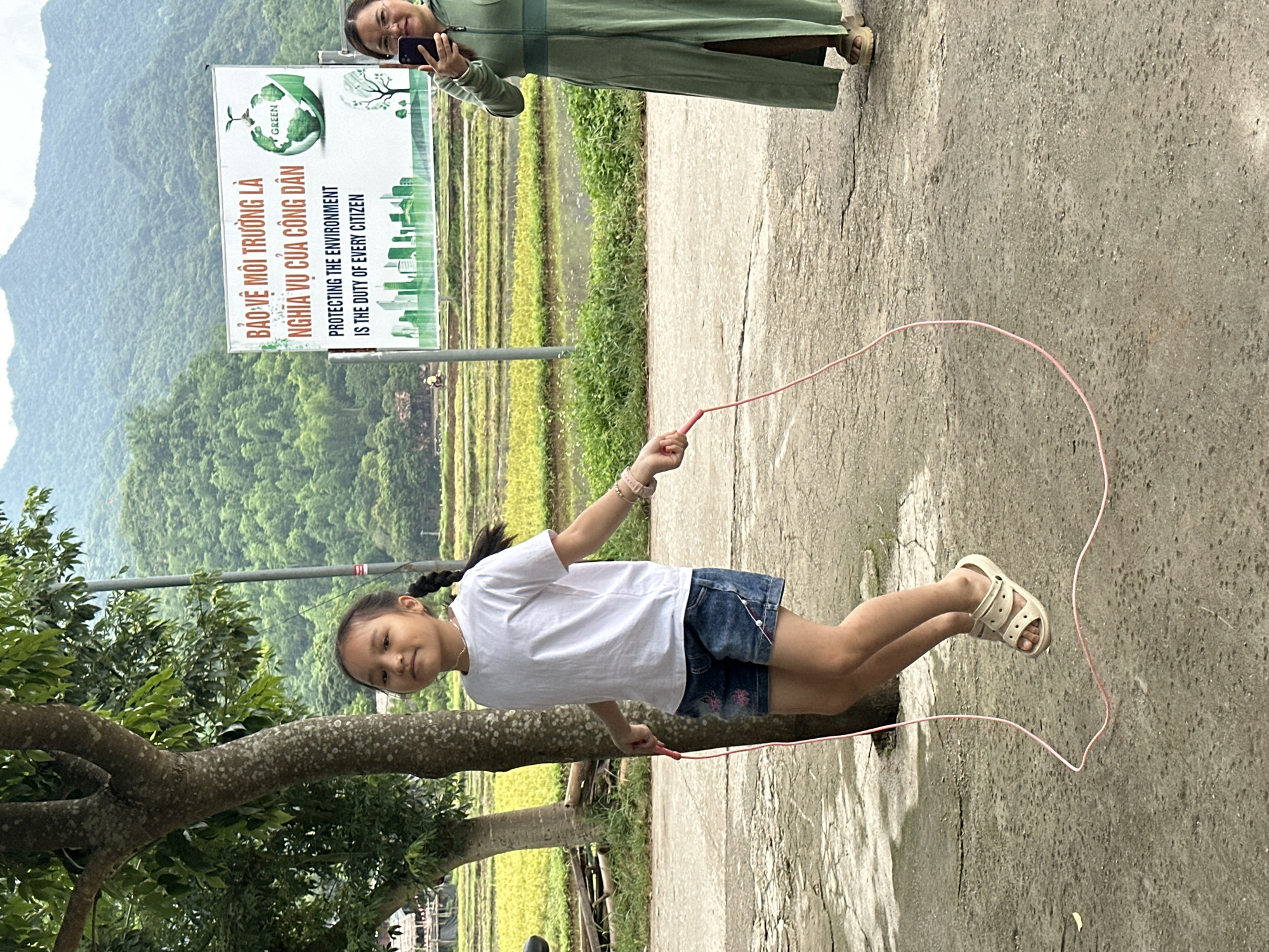 In Mai Chau, Vietnam, a young Vietnamese girl with braided hair is actively engaged in jump-roping on a concrete surface. She is wearing a white t-shirt and denim shorts, with light-colored sandals on her feet. A pink jump rope is in motion around her. To her right, a Vietnamese woman in a long, green dress is standing and holding a smartphone, appearing to take a photograph. Behind them, a sign with Vietnamese and English text regarding environmental protection is displayed, featuring a globe and cityscape graphic. The backdrop consists of lush green hills and rice paddies, indicative of a rural Vietnamese landscape.