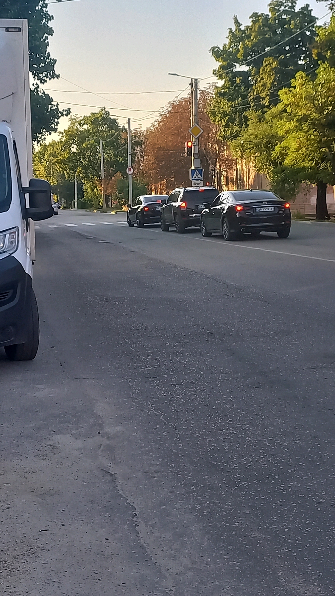 The image depicts a street scene in Kropyvnytskyi, Ukraine, with several cars stopped in traffic. In the foreground, the left side of a white van is visible. Beyond the van, a line of cars, including a black sedan and a black SUV, are stopped on the road. A traffic light is visible in the distance, showing a red light. Trees line the sides of the road, and buildings are present in the background. The sky is clear and bright, suggesting it is daytime. The road appears to be asphalt with visible cracks and some texture.