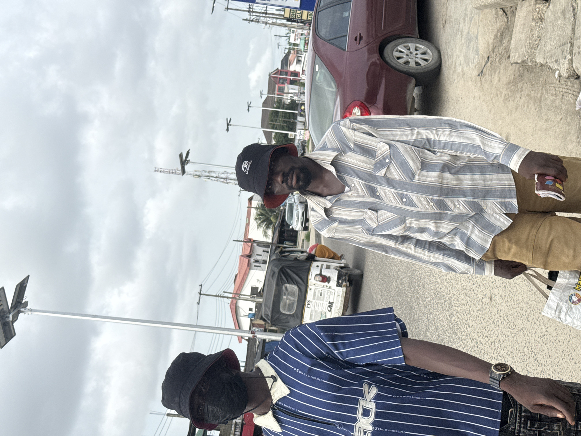 A street scene in Ado, Nigeria is captured, showcasing two individuals in the foreground. The first person, partially visible, wears a dark blue pinstripe shirt featuring a white collar, a black bucket hat with a red lining, a wristwatch, and a black face mask. The second person, more prominent, sports a light blue and white pinstripe shirt, khaki pants, and a black bucket hat with a white logo. The first is carrying what appears to be a shopping bag. A maroon car is partially visible on the right side of the frame. In the background, there is a white tuk-tuk, various buildings, telephone poles, and an overcast sky. A light pole stands to the left.